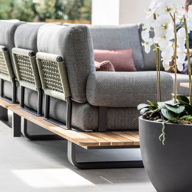 The Bay Corner Group Set with 4 Waist Pillows and a Teak Table in Olive Green, featuring gray cushions and accented with Olive Green acrylic rope, sits beside a large black planter with white orchids against a blurred background.