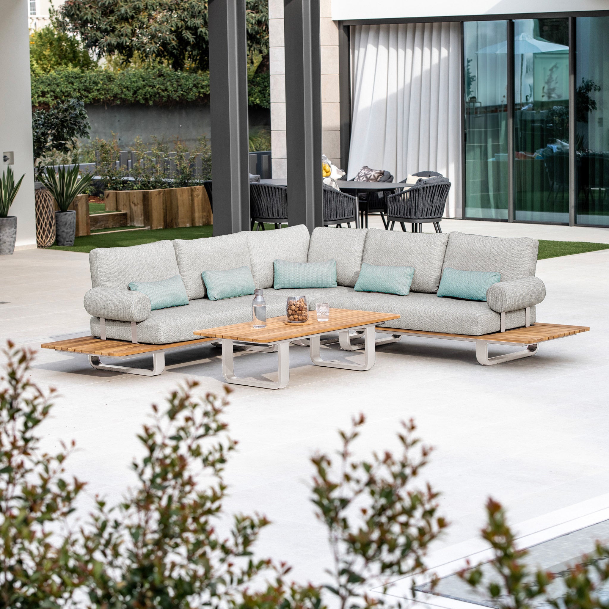 Outdoor patio featuring the Bay Corner Group Set with 4 Waist Pillows, showcasing a modern sectional sofa with a powder-coated aluminum frame, accompanied by a teak table in Latte, complete with a glass of water and snacks on the table.