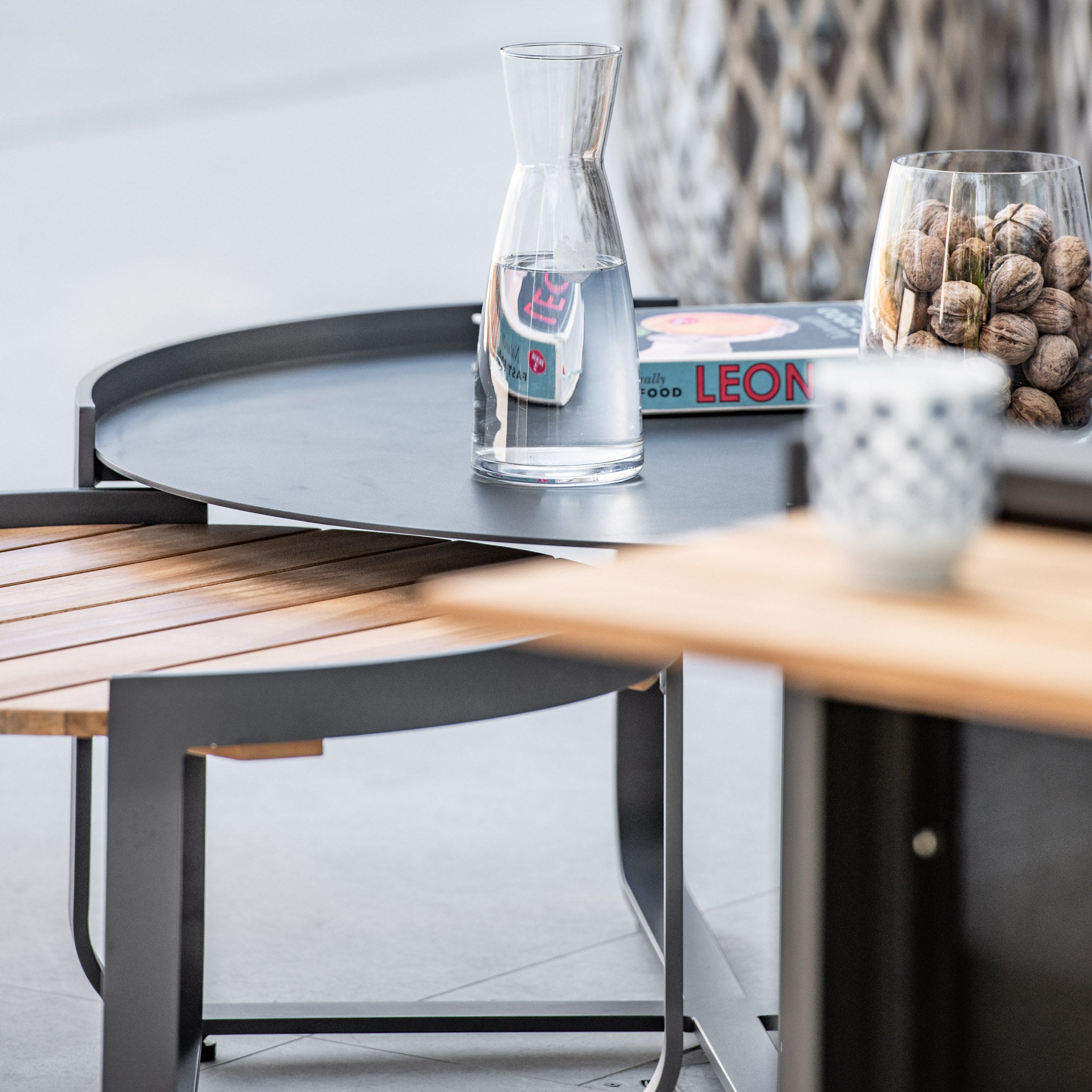 Glass pitcher on a round black metal **Bloom Coffee Table 80cm with Ceramic Top in Charcoal** with a wooden seat, a bowl of nuts, and a book named "LEON" in the background.