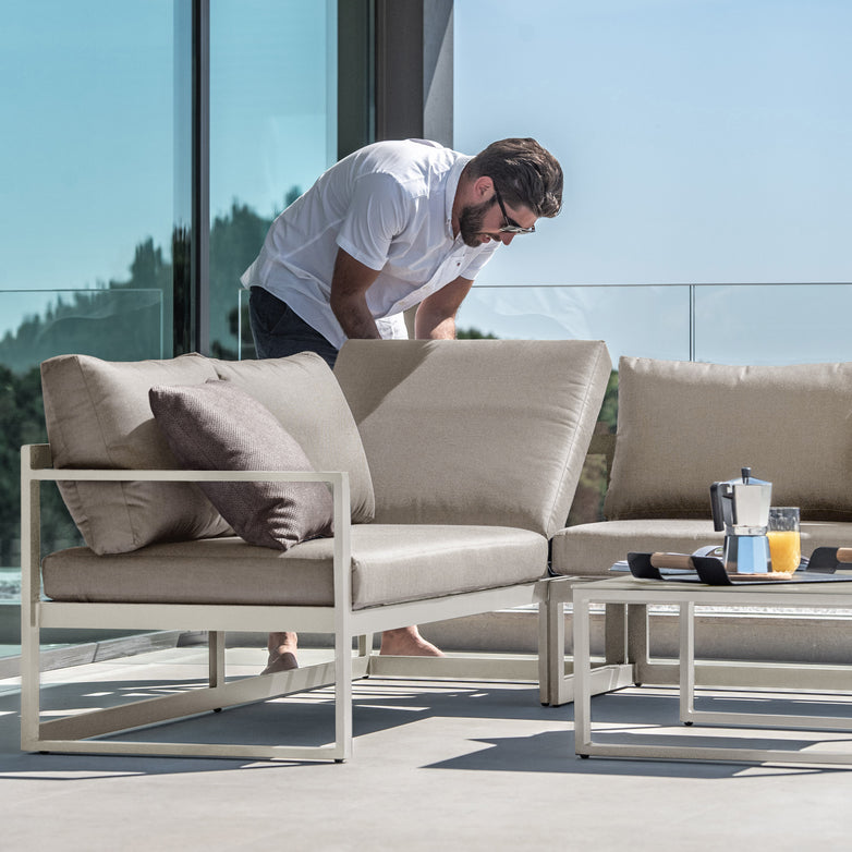 A man adjusts cushions on an outdoor sectional sofa from the Havana Corner Group Set with Reclining Feature and Coffee Table in Latte, with a glass of juice and coffee on a table beside him, enjoying his well-designed outdoor living space.