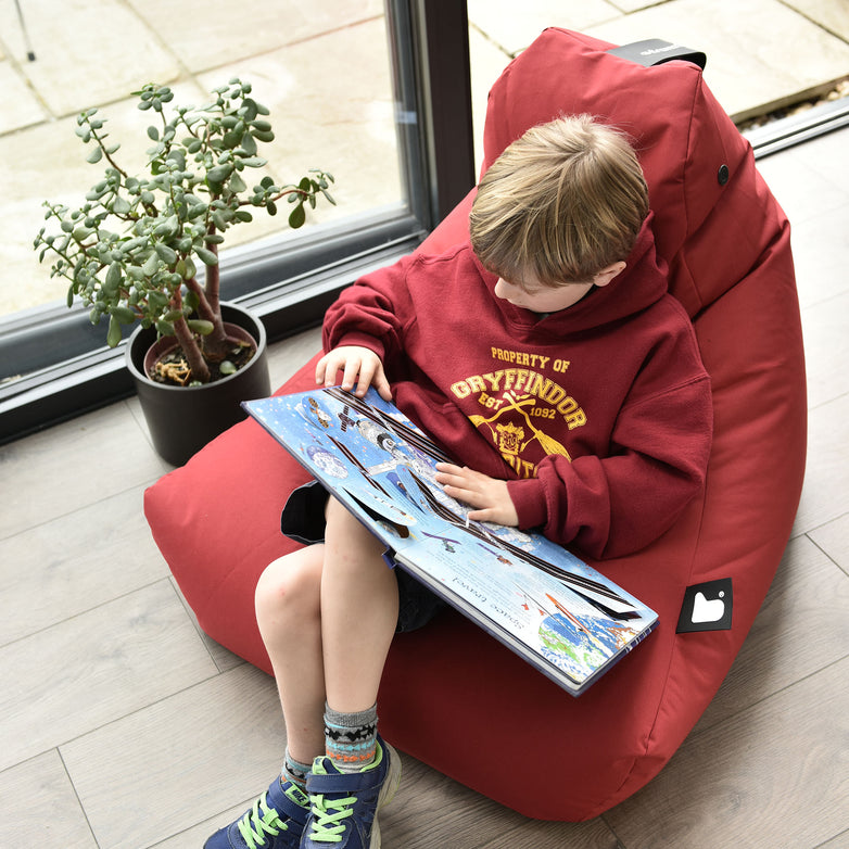 A boy reading a book while sitting on an Outdoor Mini B-Bag in Red, made of fade-resistant fabric, next to a potted plant.