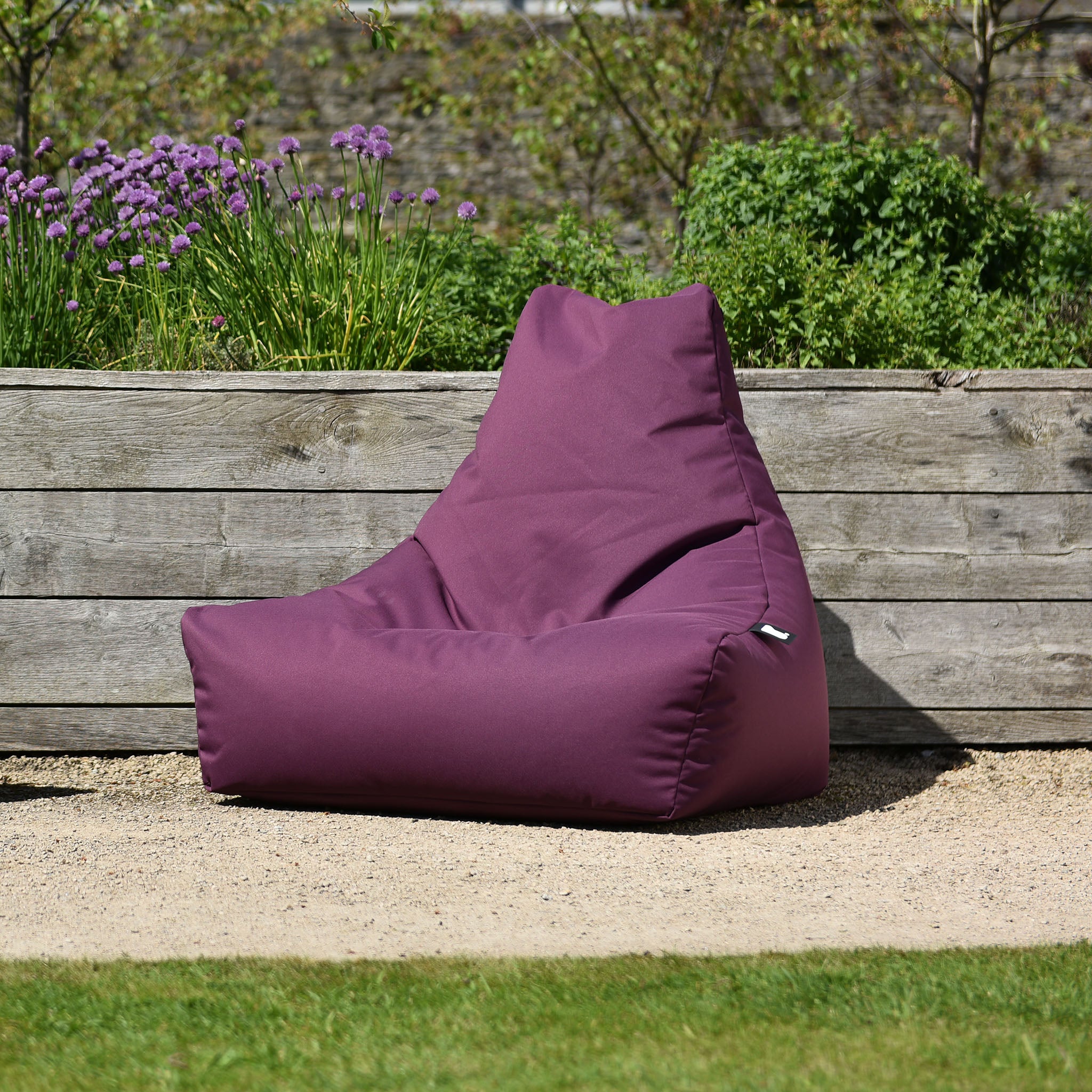 Outdoor Mighty B-Bag in Berry featuring a purple, fade-resistant fabric, placed on a sandy surface with a wooden planter and greenery in the background.