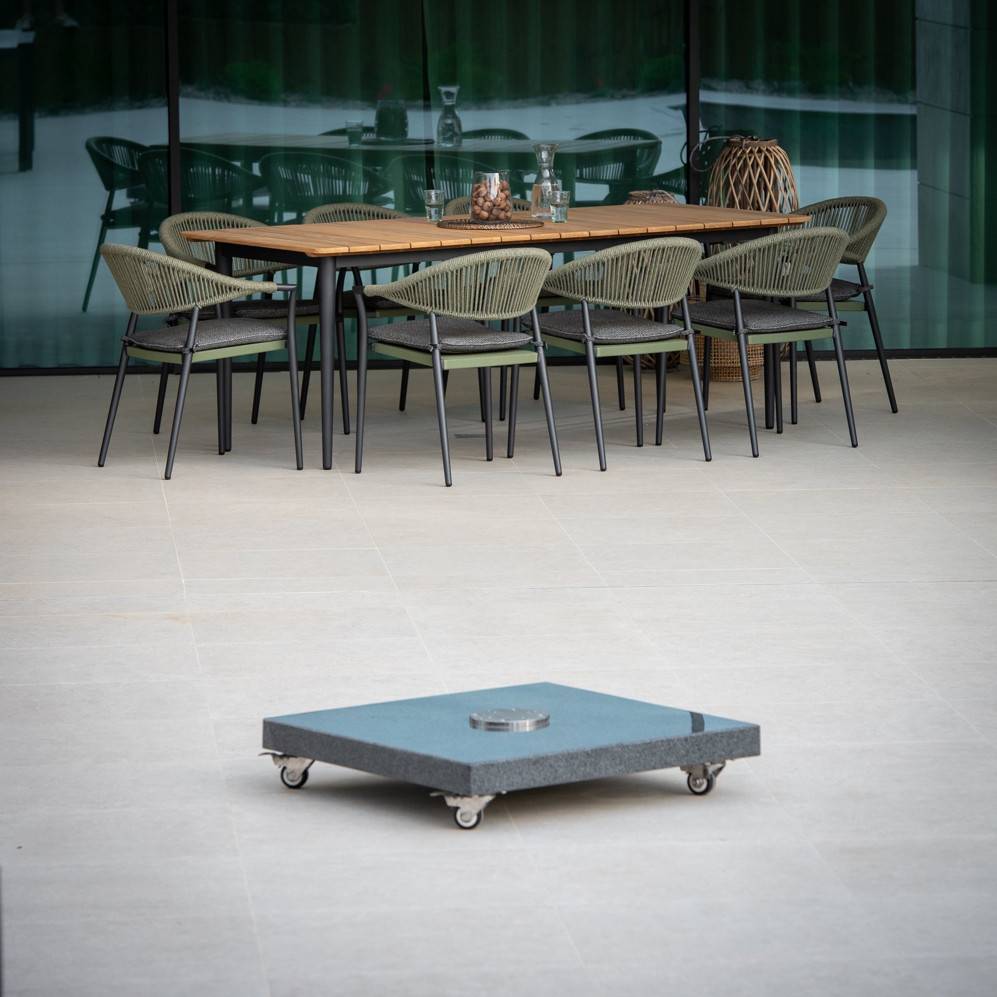 Outdoor dining area featuring a wooden table and wicker chairs. In the foreground, a square Granite 90kg Cantilever Parasol Base With Wheels.