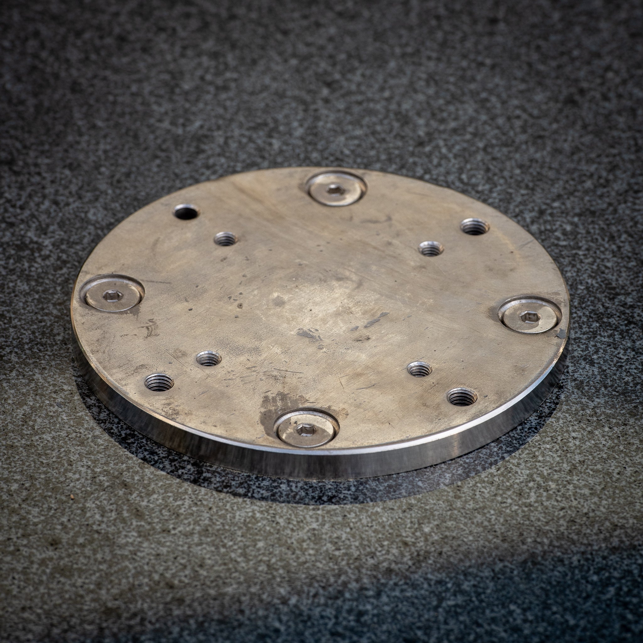 A circular metal plate with multiple holes, reminiscent of a Granite 90kg Cantilever Parasol Base With Wheels, sits elegantly on a textured gray surface.