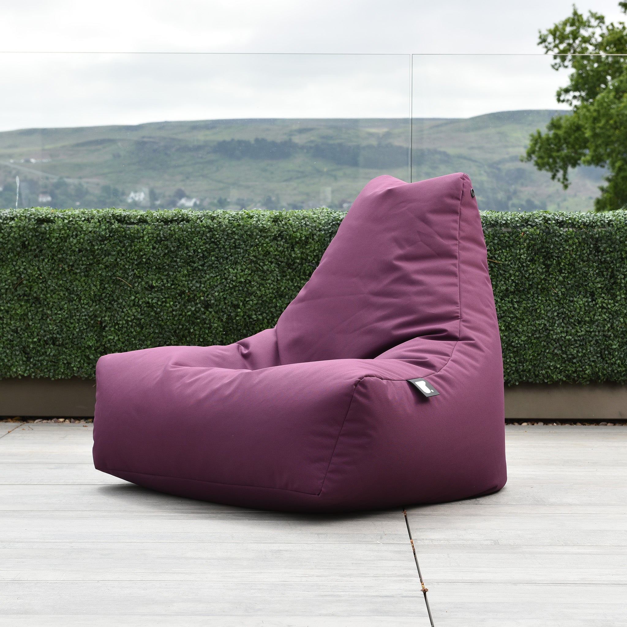 An Outdoor Mighty B-Bag in Berry, made of fade-resistant fabric, sits on an outdoor patio with a green hedge and rolling hills in the background.