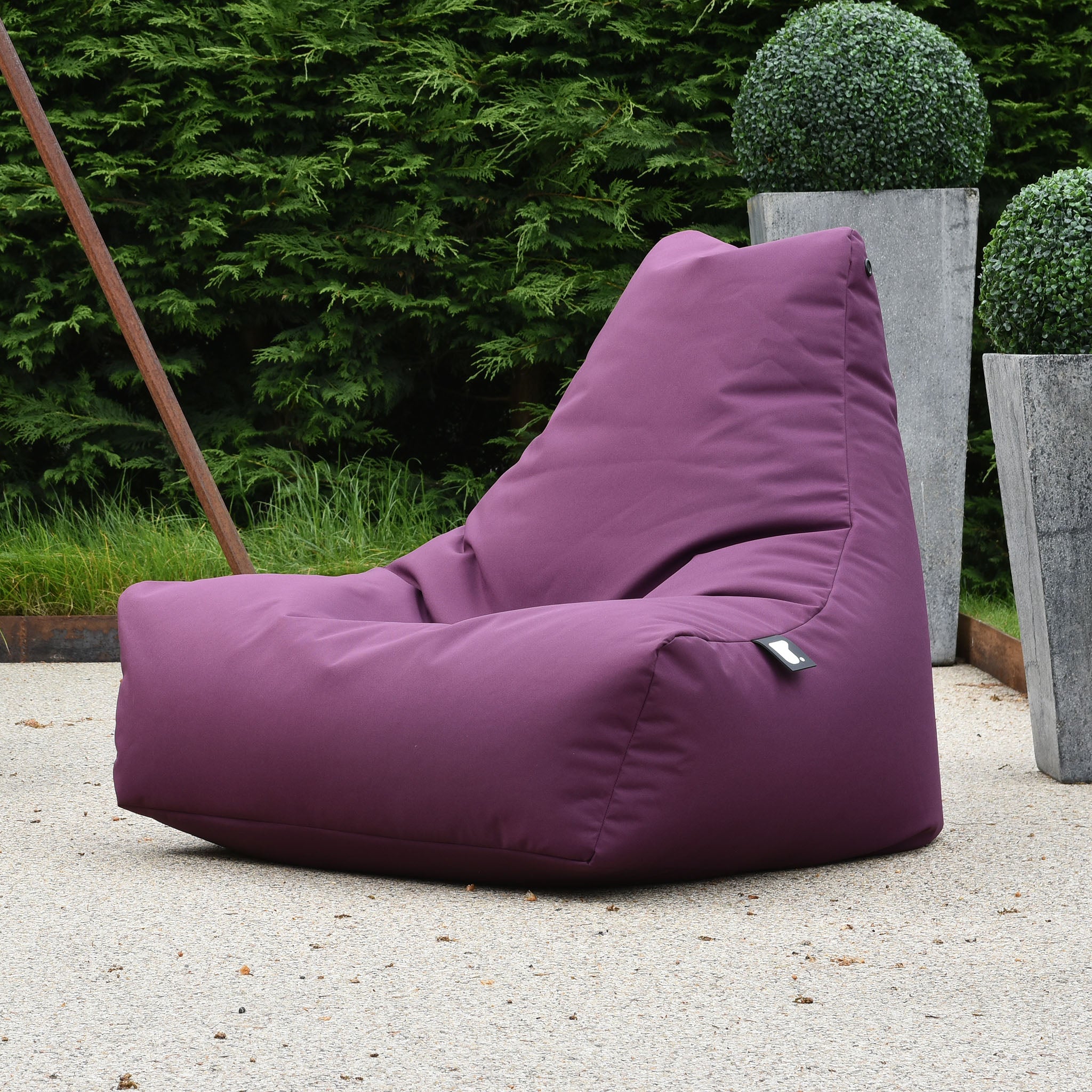 The Outdoor Mighty B-Bag in Berry graces the patio, made from fade-resistant fabric, nestled among green bushes and decorative planters in the background.