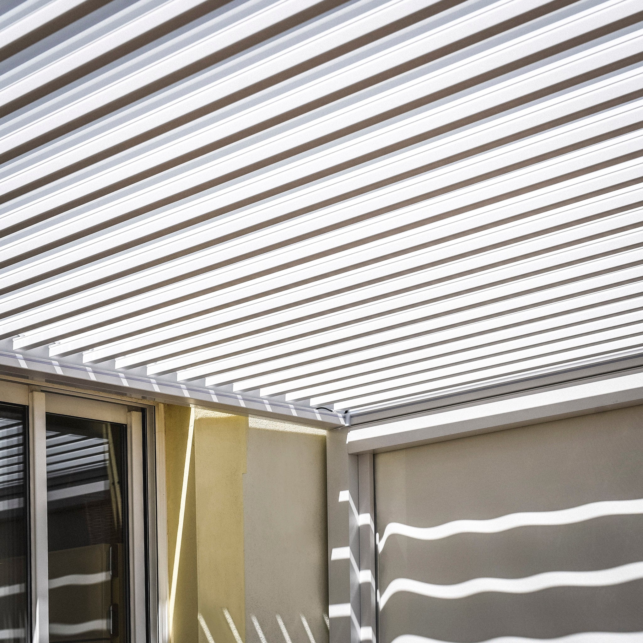 The white horizontal slatted roof of the PergoSTET 3m x 3m Square Pergola with 3 Drop Sides and LED Lighting creates striped shadows on a wall and glass door below.