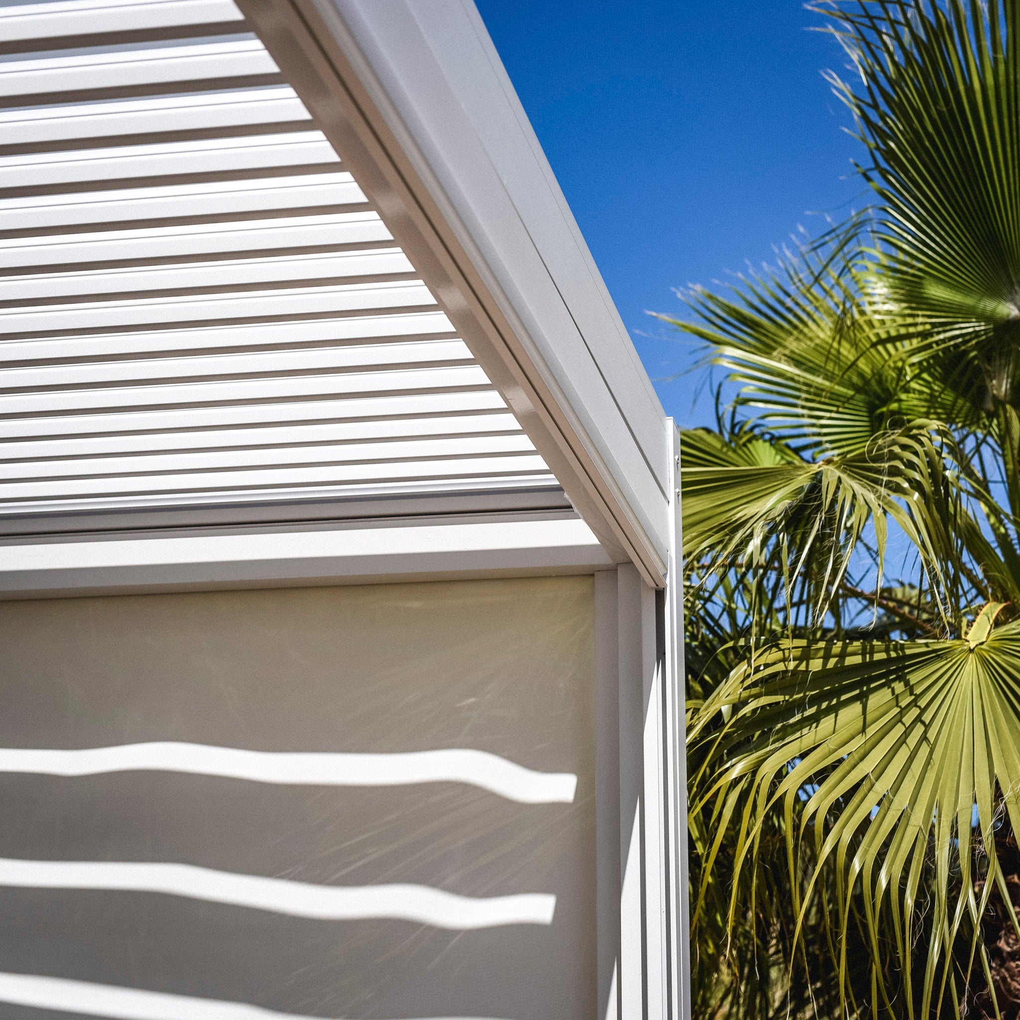 The PergoSTET 3m x 3m Square Pergola with 3 Drop Sides and LED Lighting in White casting shadows, with a palm tree and clear blue sky in the background, features a built-in drainage system for modern convenience.