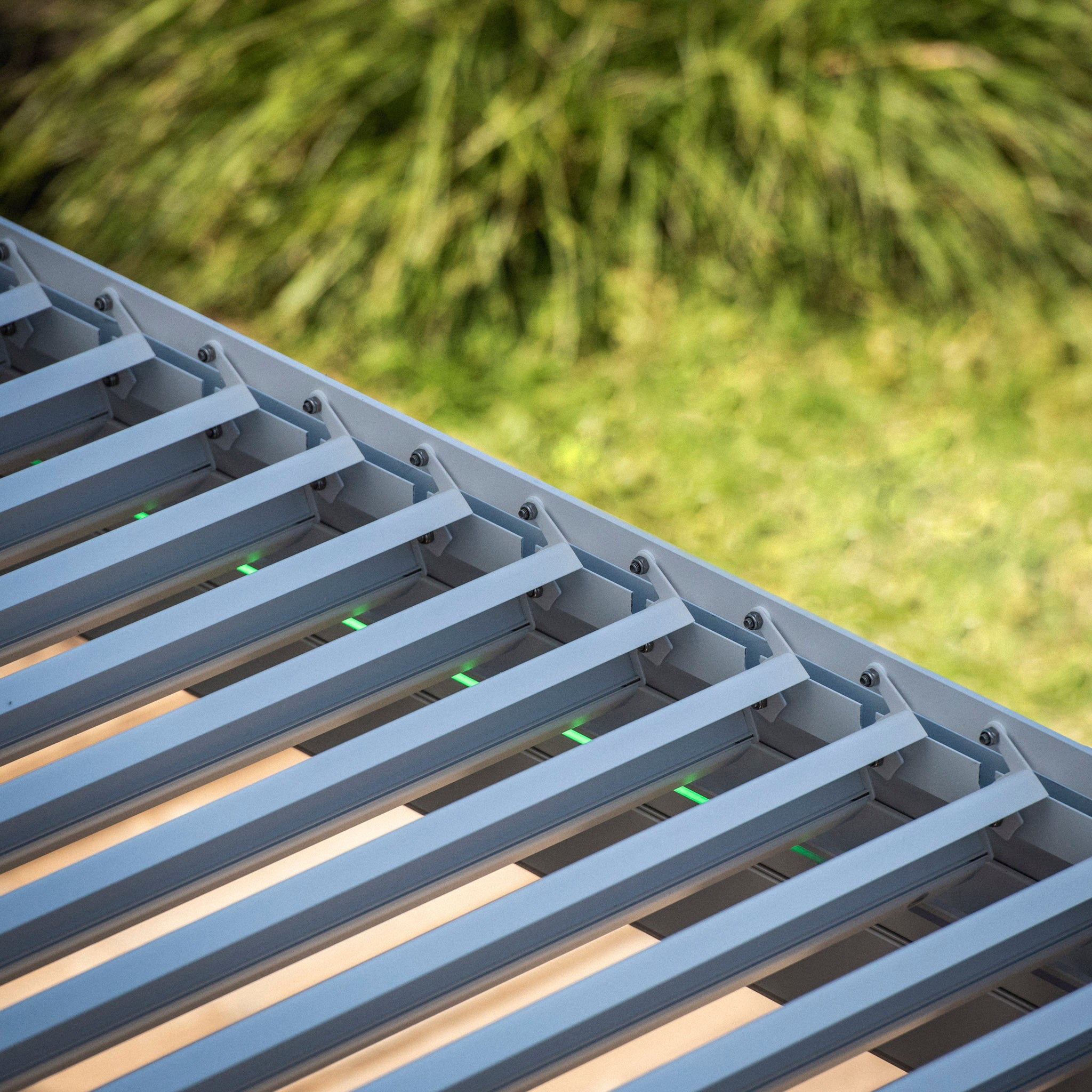 Close-up of a white slatted structure, possibly the PergoSTET 3m x 3m Square Pergola with 3 Drop Sides and LED Lighting, with a green, blurry background of foliage in a garden setting.