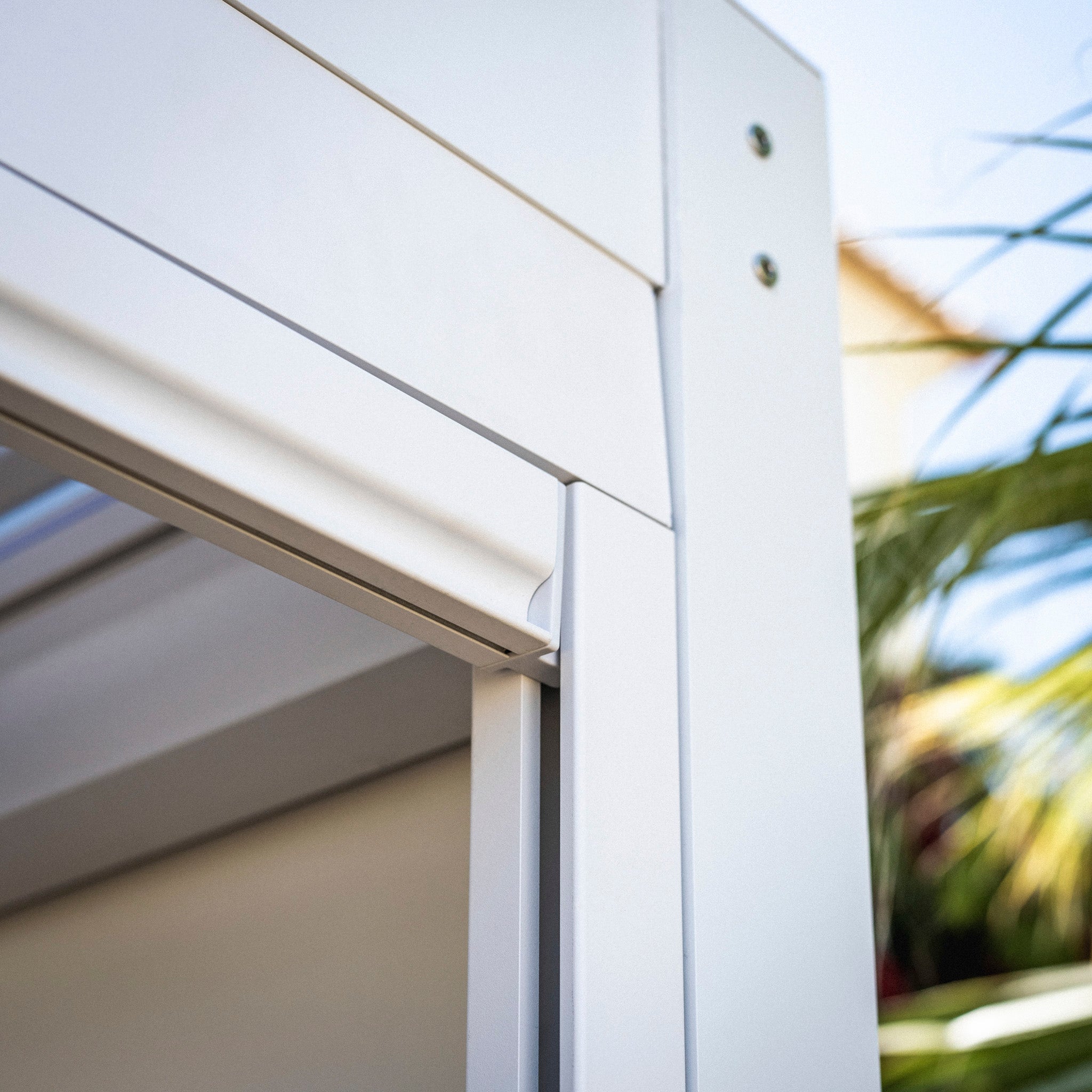 Close-up of a sleek, white modern window frame with a blue sky and green palm leaves in the background, seamlessly integrated with the PergoSTET 3m x 3m Square Pergola with 3 Drop Sides and LED Lighting in White, featuring a built-in drainage system.