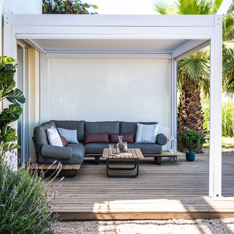 Outdoor patio with a gray sectional sofa, wooden table, and green plants surrounded by the PergoSTET 3m x 3m Square Pergola with 3 Drop Sides and LED Lighting in White, featuring a built-in drainage system.