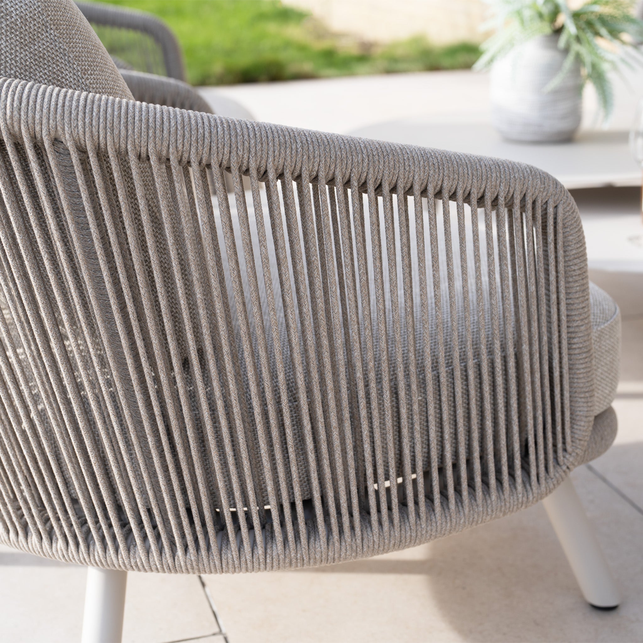 Close-up of a woven outdoor chair with an olefin rope weave and beige cushion on a concrete patio near a table and potted plant. Part of the Dekota 3 Seat Rope Sofa Set with Coffee Tables and Footstool in Fawn, this piece enhances any outdoor furniture setting.