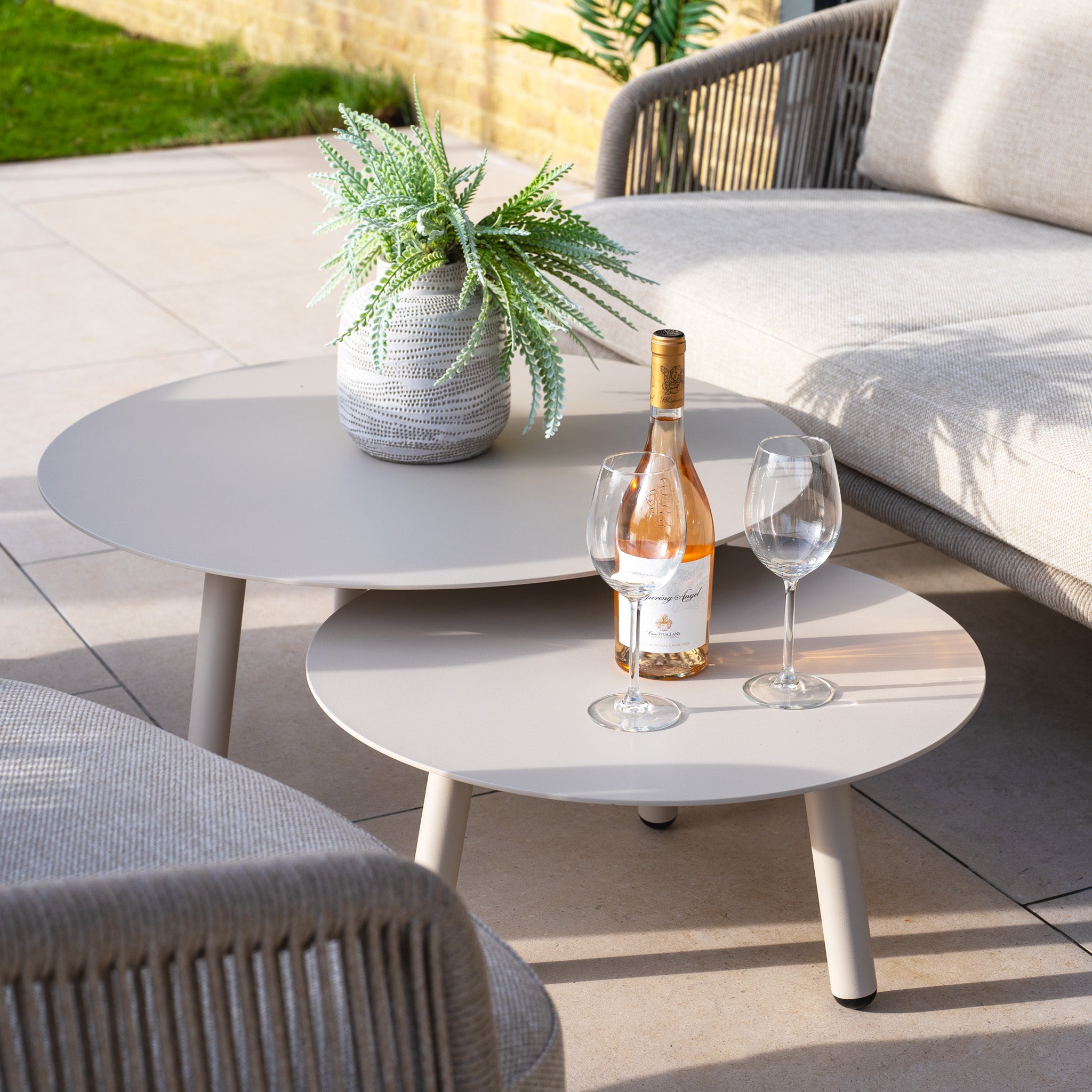 Contemporary patio setup with the Dekota 3 Seat Rope Sofa Set in Fawn, featuring coffee tables, footstool, wine bottle, glasses, and a potted plant. The wicker sofa and olefin rope weave design provide an exceptional outdoor furniture experience.