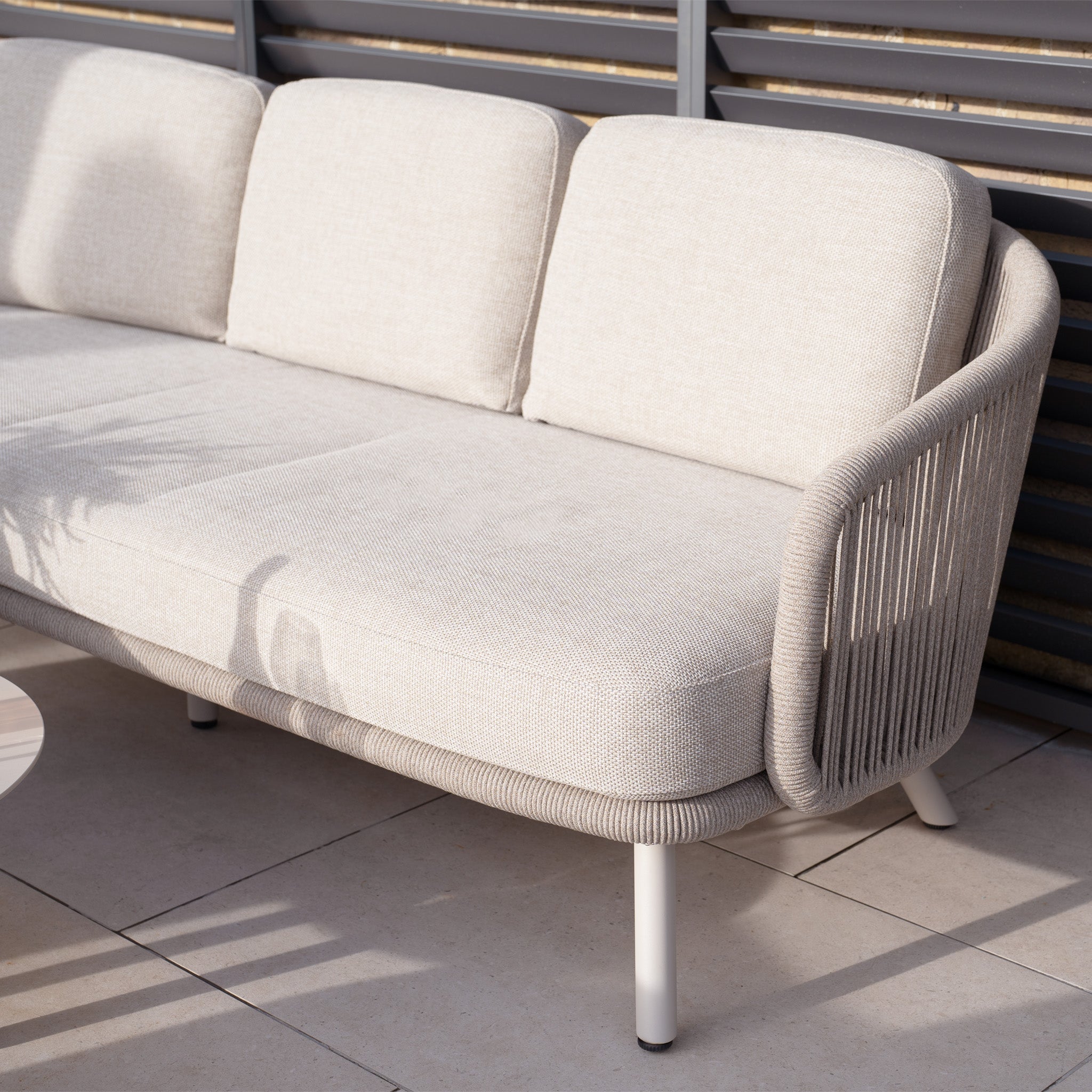 The Dekota 3 Seat Rope Sofa Set with Coffee Tables and Footstool in Fawn features a modern design with an olefin rope weave and white legs. Perfect for tiled patios, this outdoor furniture captures sunlight beautifully, casting intriguing shadows across the seating area.