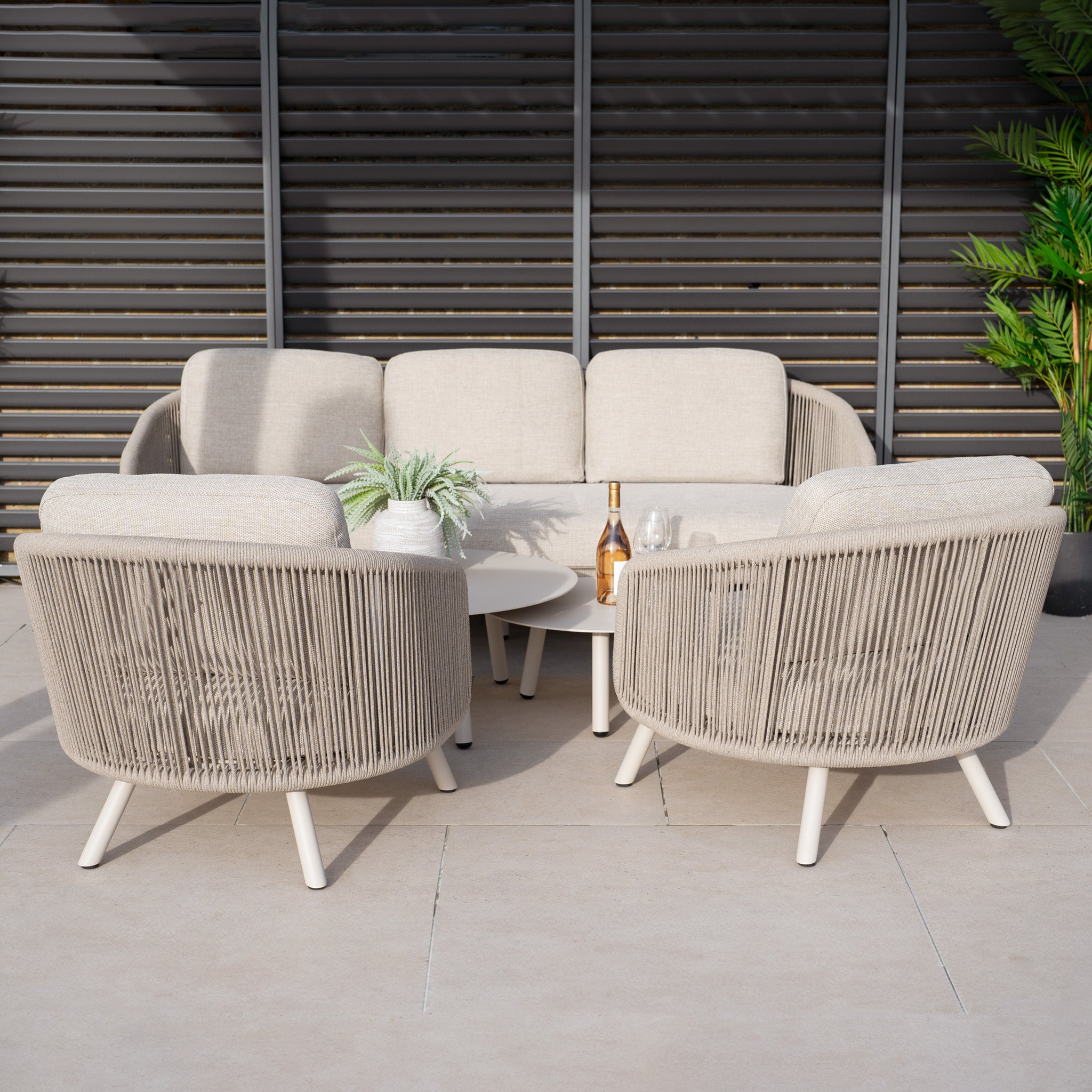 Outdoor seating with the Dekota 3 Seat Rope Sofa Set in Fawn includes cushioned chairs around a coffee table. A bottle and glasses sit on top, and greenery from a nearby potted plant complements the olefin rope weave, enhancing durability and style.