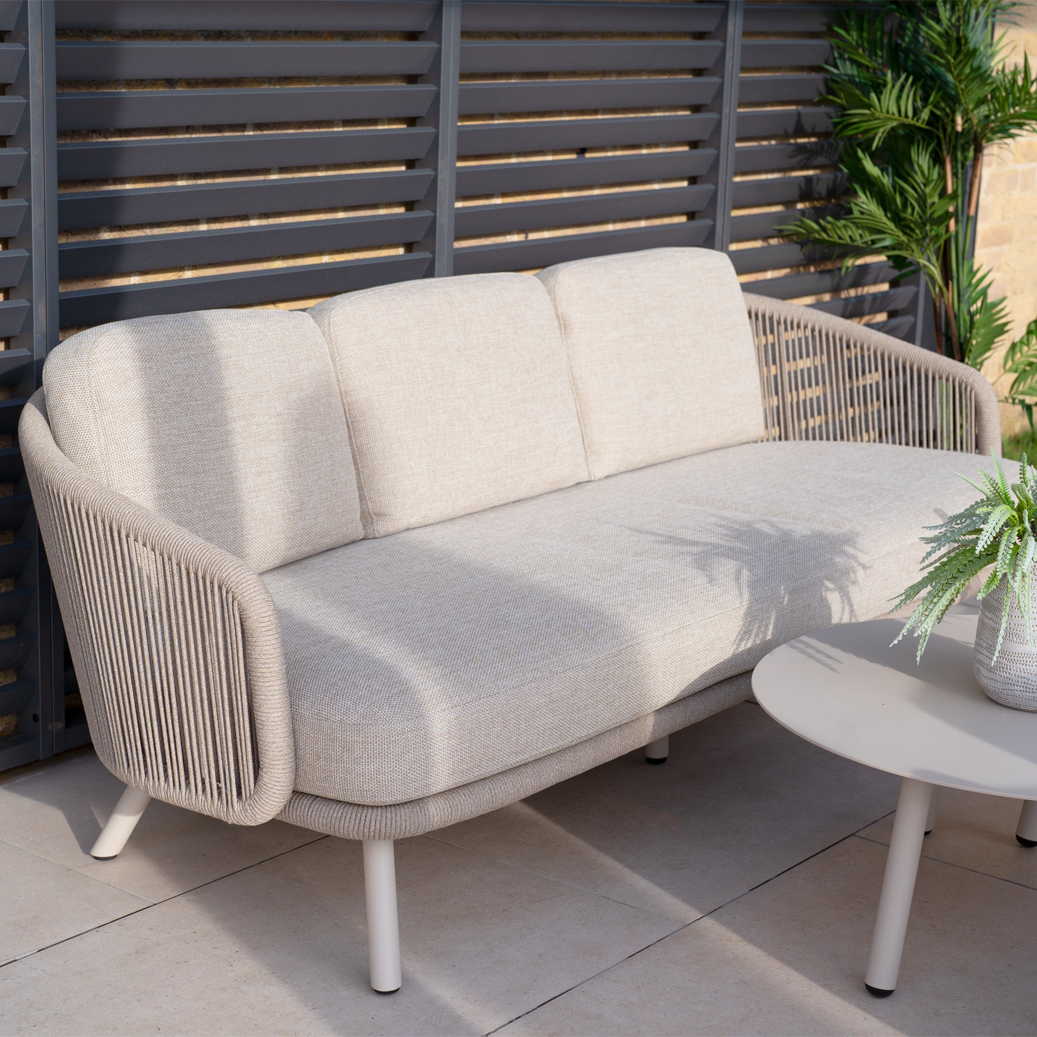 The Dekota 3 Seat Rope Sofa Set with Coffee Tables and Footstool in Fawn features a beige outdoor sofa with olefin rope weave sides and sits elegantly beside a round table topped with a potted plant, showcasing premium outdoor furniture style.