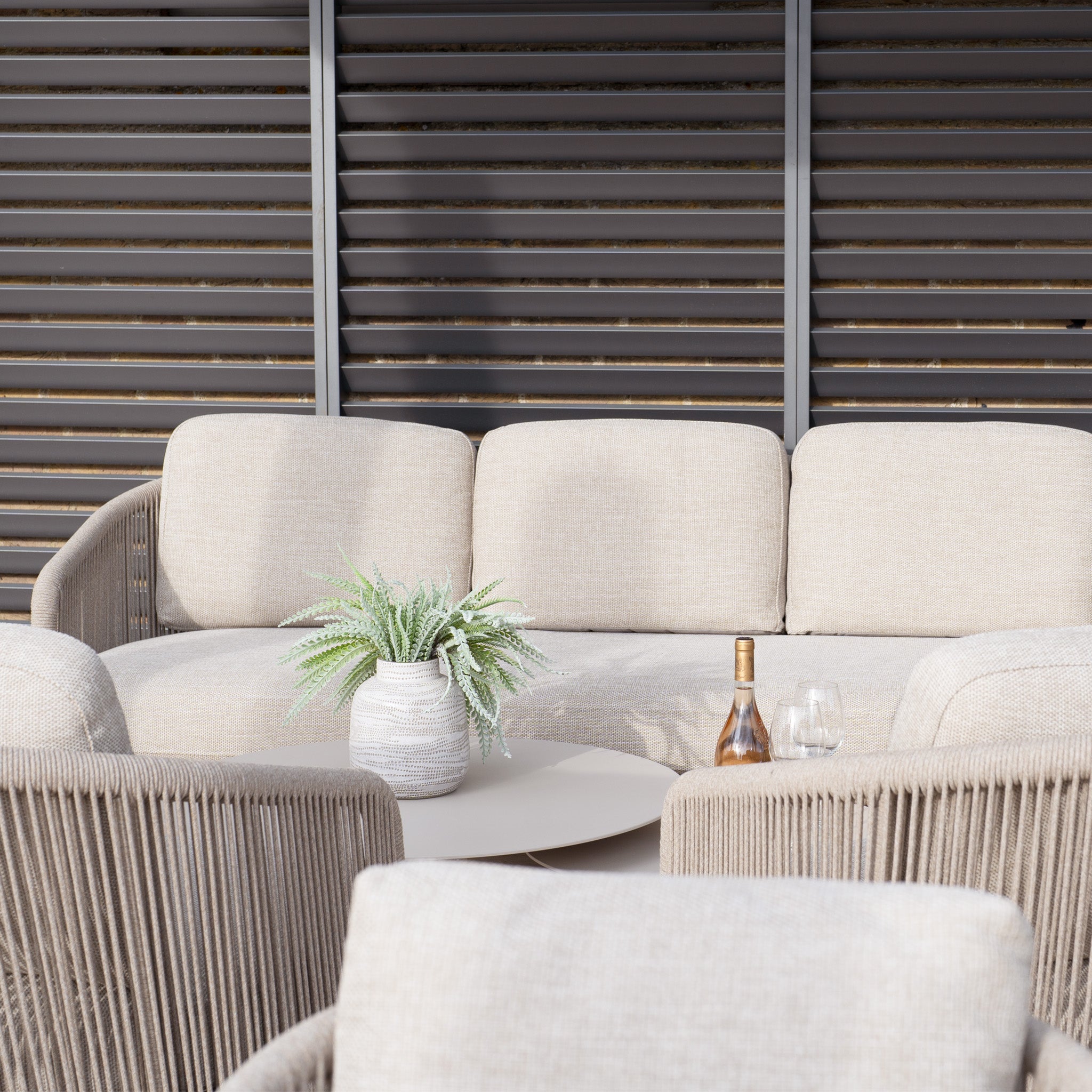 The Dekota 3 Seat Rope Sofa Set, crafted in fawn and featuring coffee tables and a footstool, includes a table with a potted plant and wine glasses. A stylish louvered wall highlights its elegant olefin rope weave.