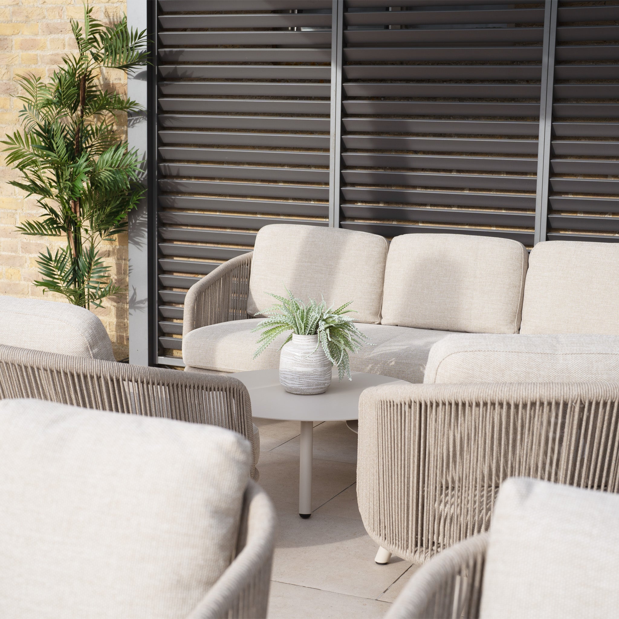 The outdoor seating area boasts the Dekota 3 Seat Rope Sofa Set with beige cushions, a footstool, and round coffee tables. The elegant set features an intricate olefin rope weave for durability and style, complemented by a potted plant.