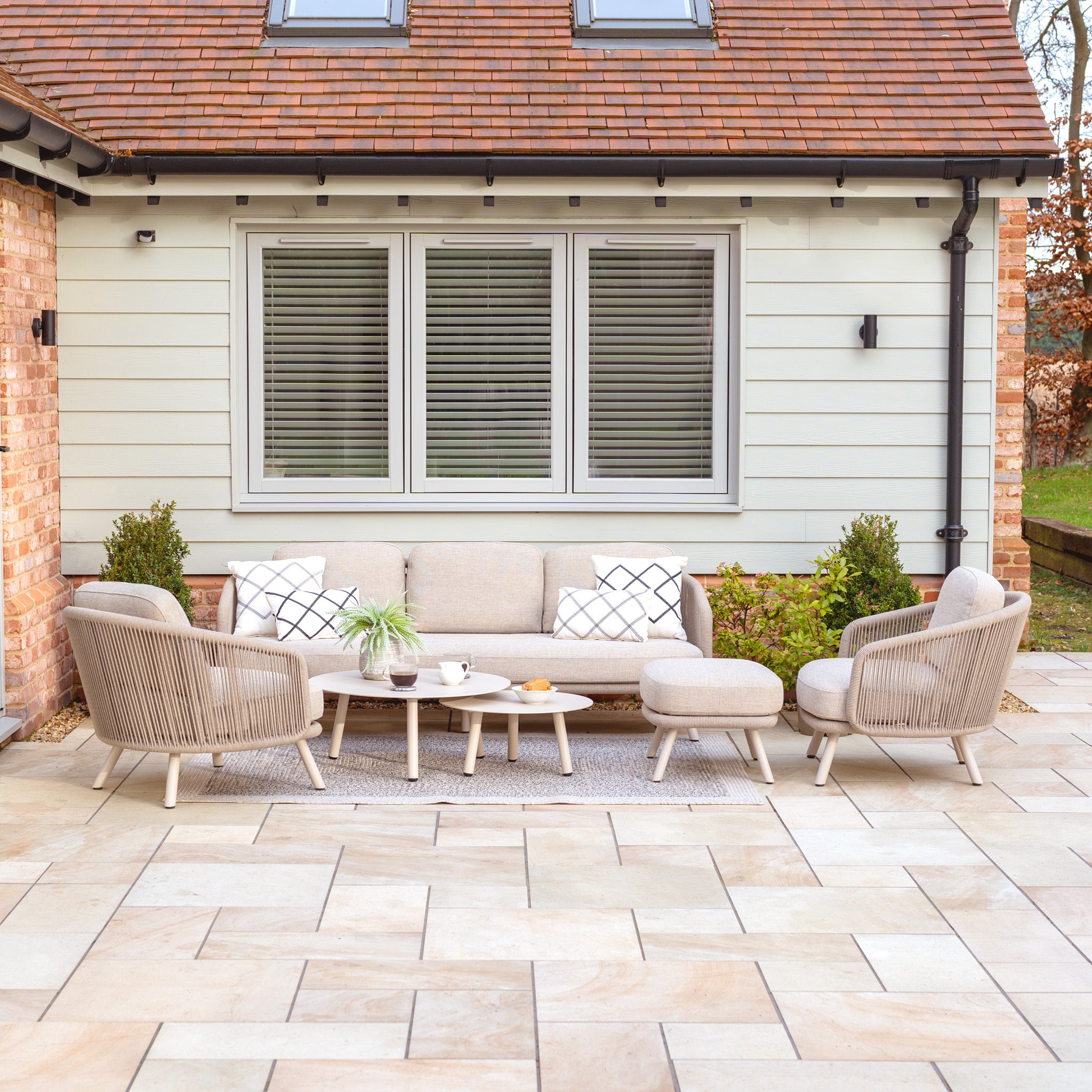 The outdoor patio highlights a Dekota 3 Seat Rope Sofa Set in Fawn, including coffee tables and a footstool, amid lush plants. Its sophisticated design pairs well with the backdrop of windows and brick details, showcasing its durable powder-coated aluminium frame for lasting elegance.