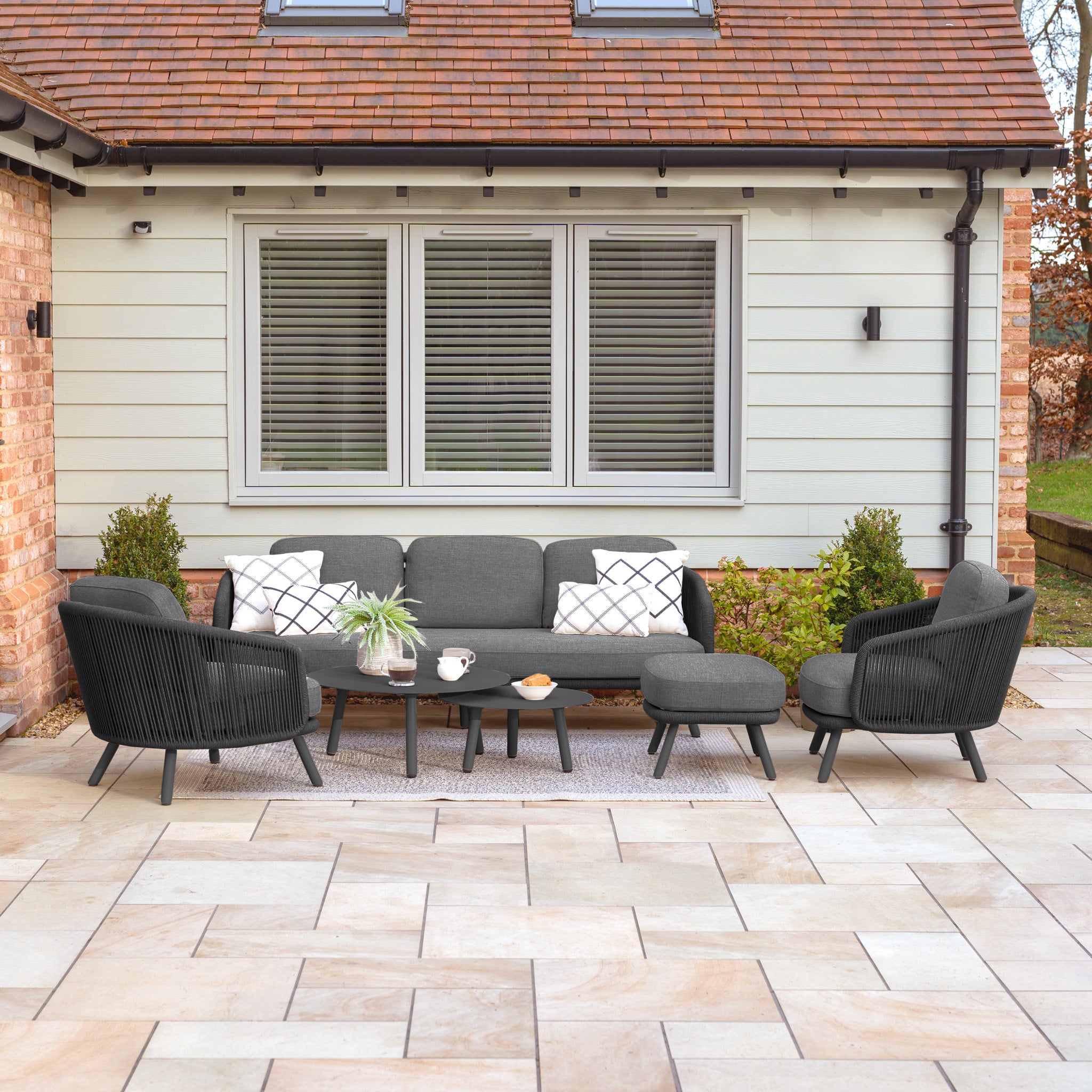 The patio showcases the stylish Dekota 3 Seat Rope Sofa Set in charcoal, complete with coffee tables and a footstool, surrounded by vibrant plants and decorative olefin pillows, all in front of a house featuring charming windows and shutters.