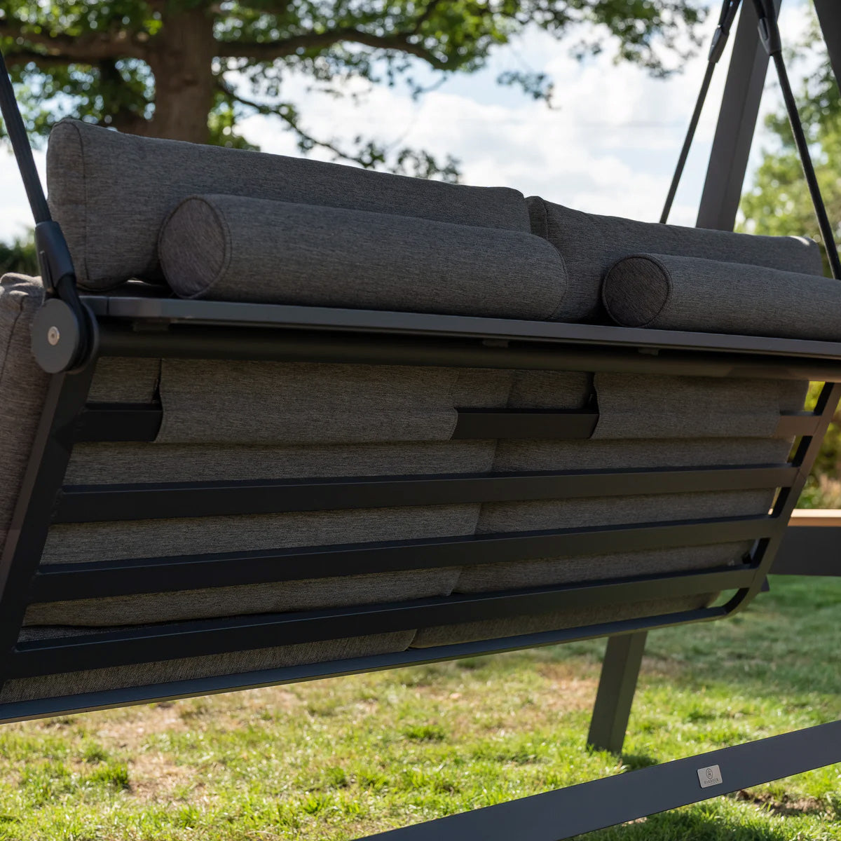 A close-up of a charcoal cushioned outdoor swing on a powder-coated aluminium frame in a garden with a tree in the background. The Lima Deluxe Aluminium Swing Seat in Charcoal features an Olefin canopy for added comfort and style.