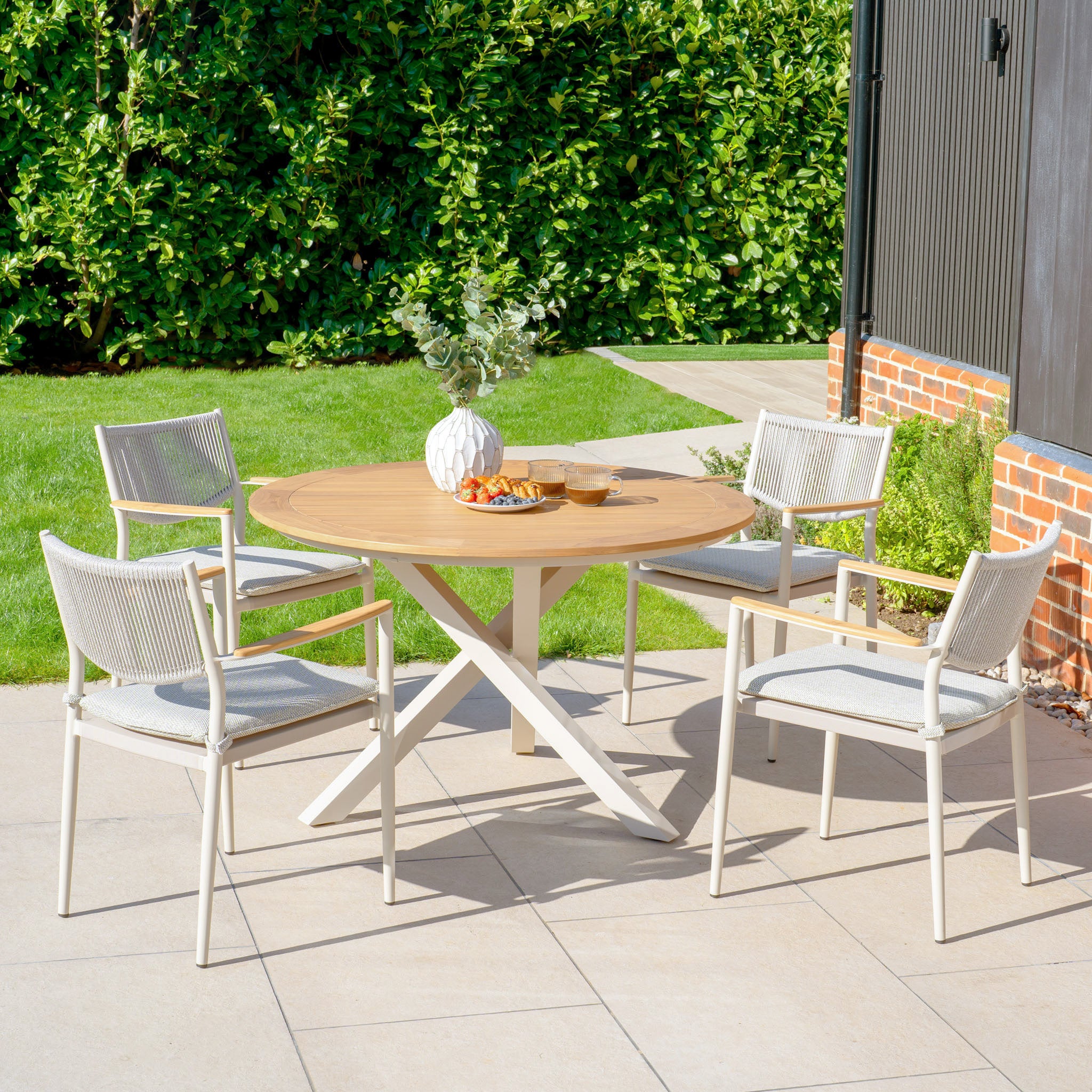 The outdoor patio showcases the Tellaro 4 Seat Round Dining Set with a teak table in Latte, complemented by four chairs crafted with weather-resistant rope, all beautifully placed on a stone terrace amid lush greenery.