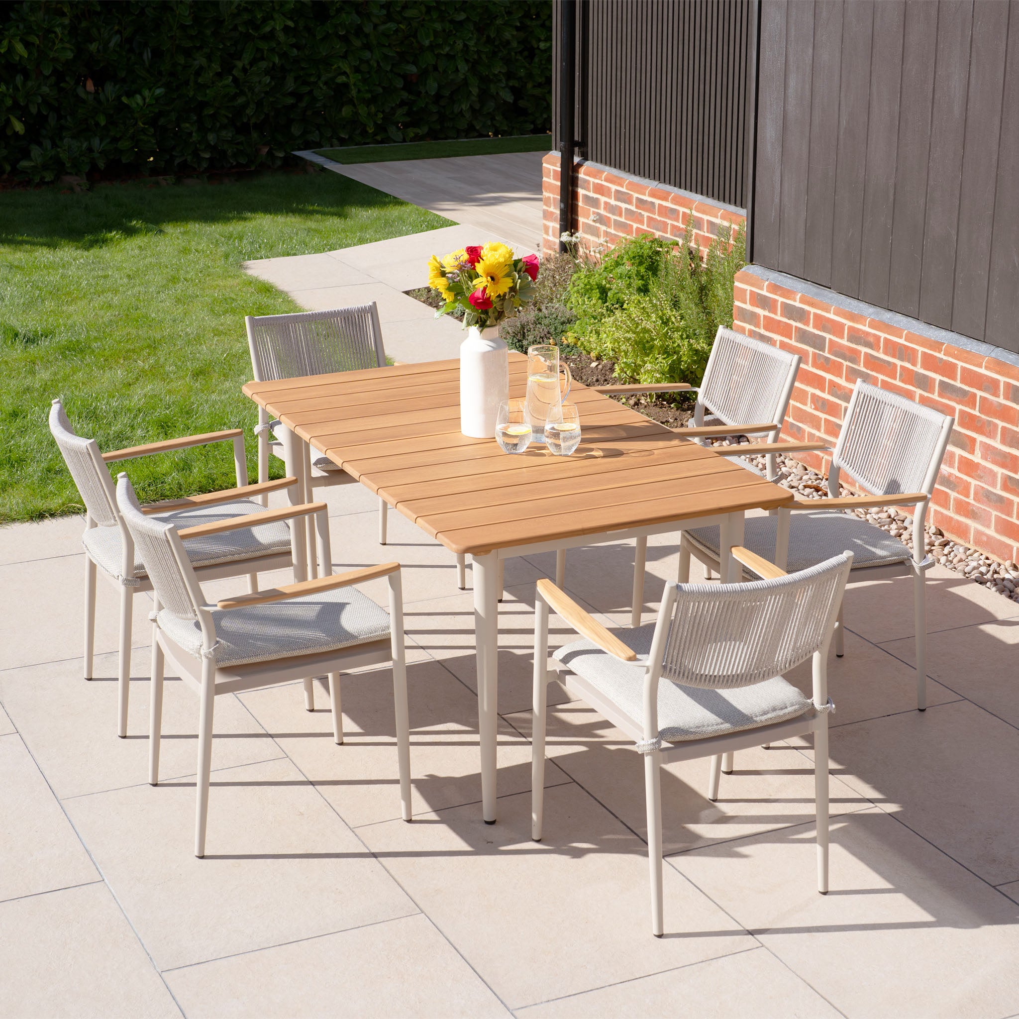 Experience outdoor living at its best with the Tellaro 6 Seat Rectangular Dining Set in Latte, featuring a teak table and six chairs. Surrounded by vibrant flowers, sunlight, lush grass, and a charming brick wall, it creates an inviting ambiance.