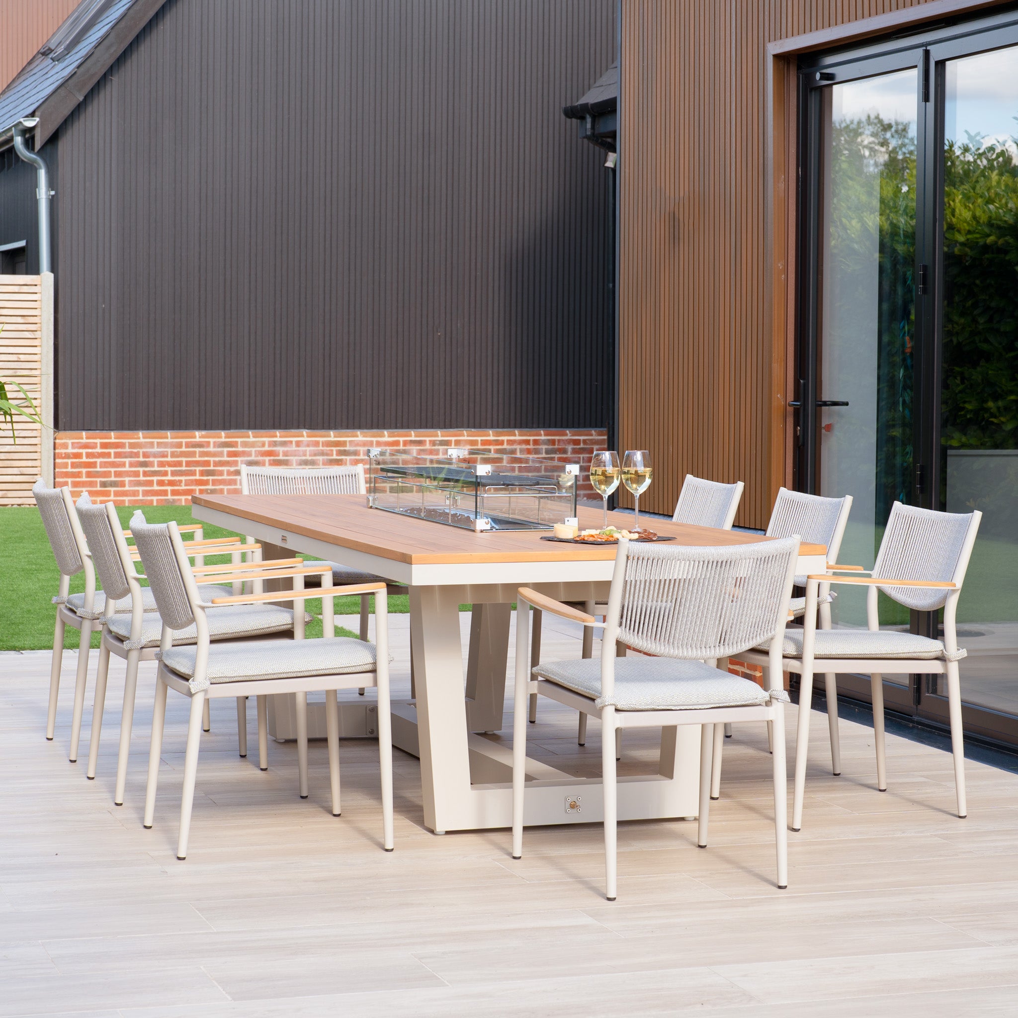 In an outdoor dining space, a Tellaro 8 Seat Rectangular Teak Firepit Dining Set in Latte is arranged with eight chairs surrounding a wooden table, beautifully set off by two glasses of white wine. The contemporary architecture's elegant style is enhanced by the powder-coated aluminium details, achieving an ideal mix of comfort and sophistication.