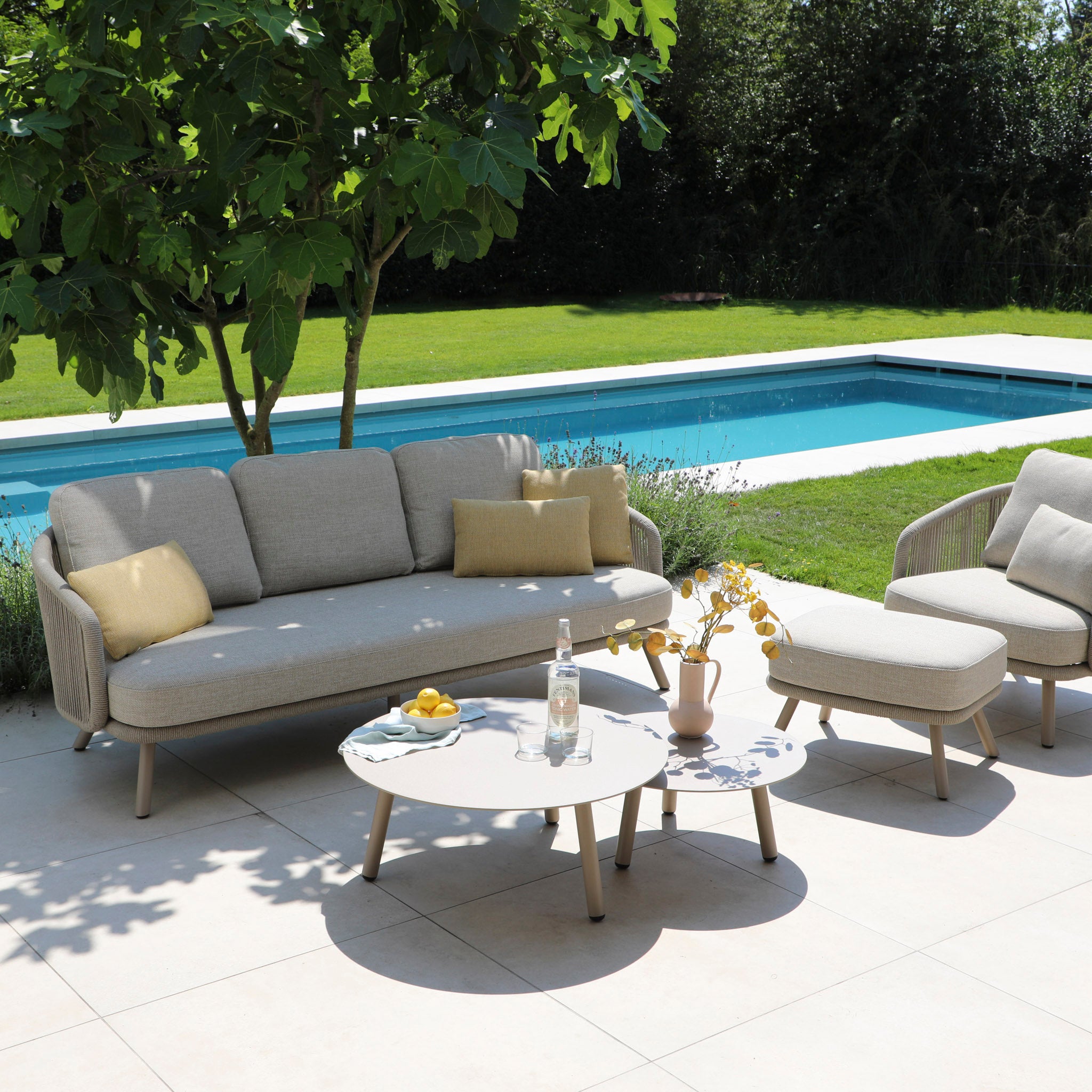 Enjoy an outdoor patio with the Dekota 3 Seat Rope Sofa Set in Fawn, featuring a hydrophobic olefin rope weave and beige cushions, complemented by a round table. Lush plants enhance the ambiance against a shimmering pool backdrop, with a durable powder-coated aluminum frame.