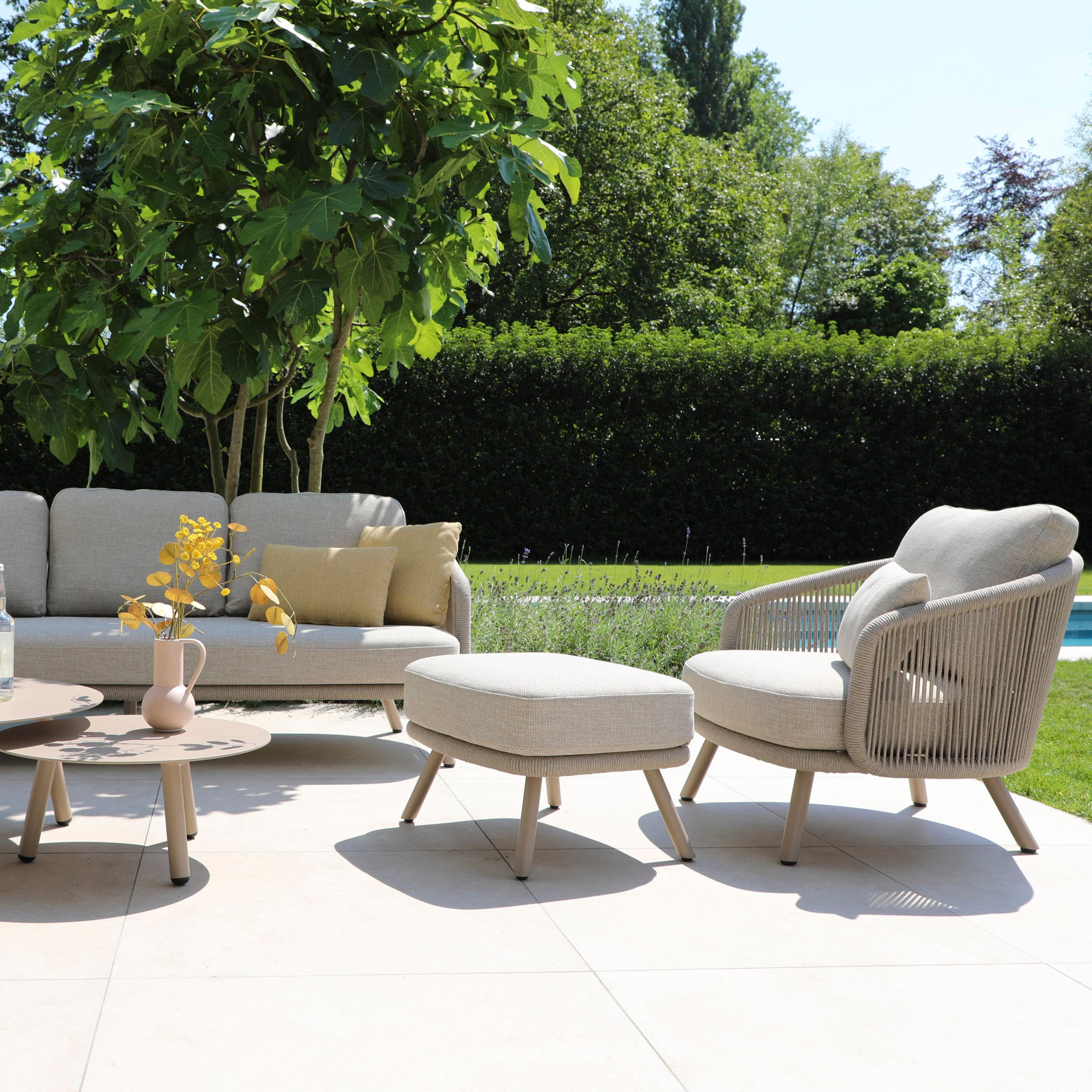 An outdoor patio showcases the Dekota 3 Seat Rope Sofa Set in Fawn, featuring a sturdy aluminum frame with beige cushions. The hydrophobic olefin rope weave complements the included coffee tables and footstool, set amidst lush greenery and a shimmering swimming pool.