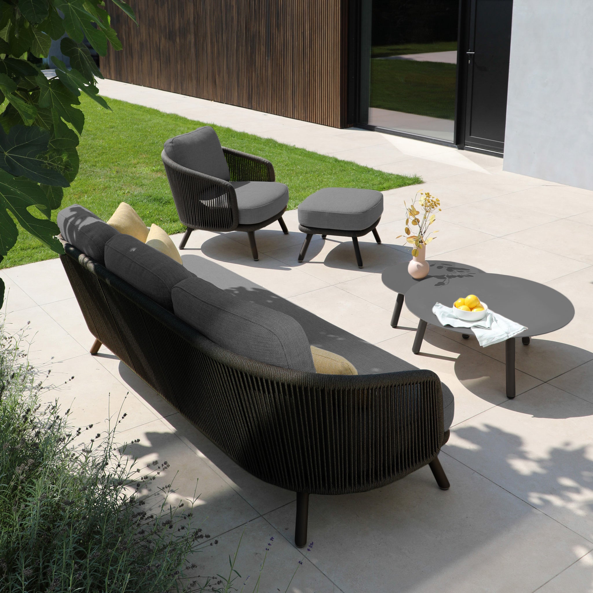 Outdoor patio featuring the Dekota 3 Seat Rope Sofa Set with Coffee Tables and Footstool in charcoal, crafted with luxurious olefin fabric. A round table showcases a vase and fruit bowl, nestled near a lush green lawn and vibrant plants for ultimate outdoor luxury.