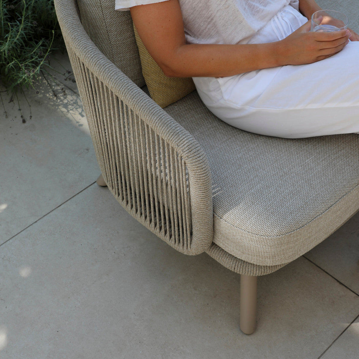 On a sunny patio, a person relaxes on the fawn Dekota 3 Seat Aluminium Sofa Set with Coffee Tables and Footstool, sipping a drink. Surrounded by hydrophobic olefin rope weave and tiles, they embrace serene outdoor comfort.