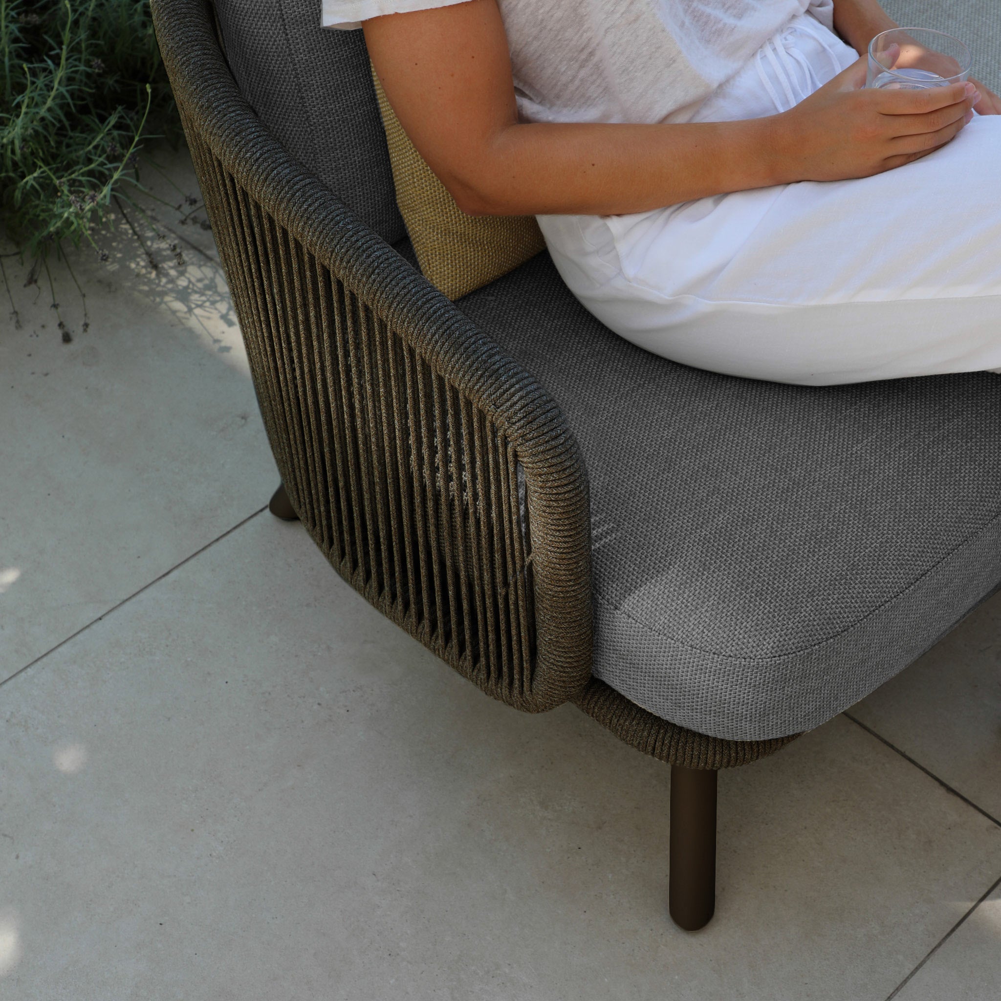 A person in white relaxes with a glass in hand on a gray cushioned chair from the Dekota 3 Seat Rope Sofa Set, featuring charcoal olefin fabric that adds luxury to the serene outdoor setting.