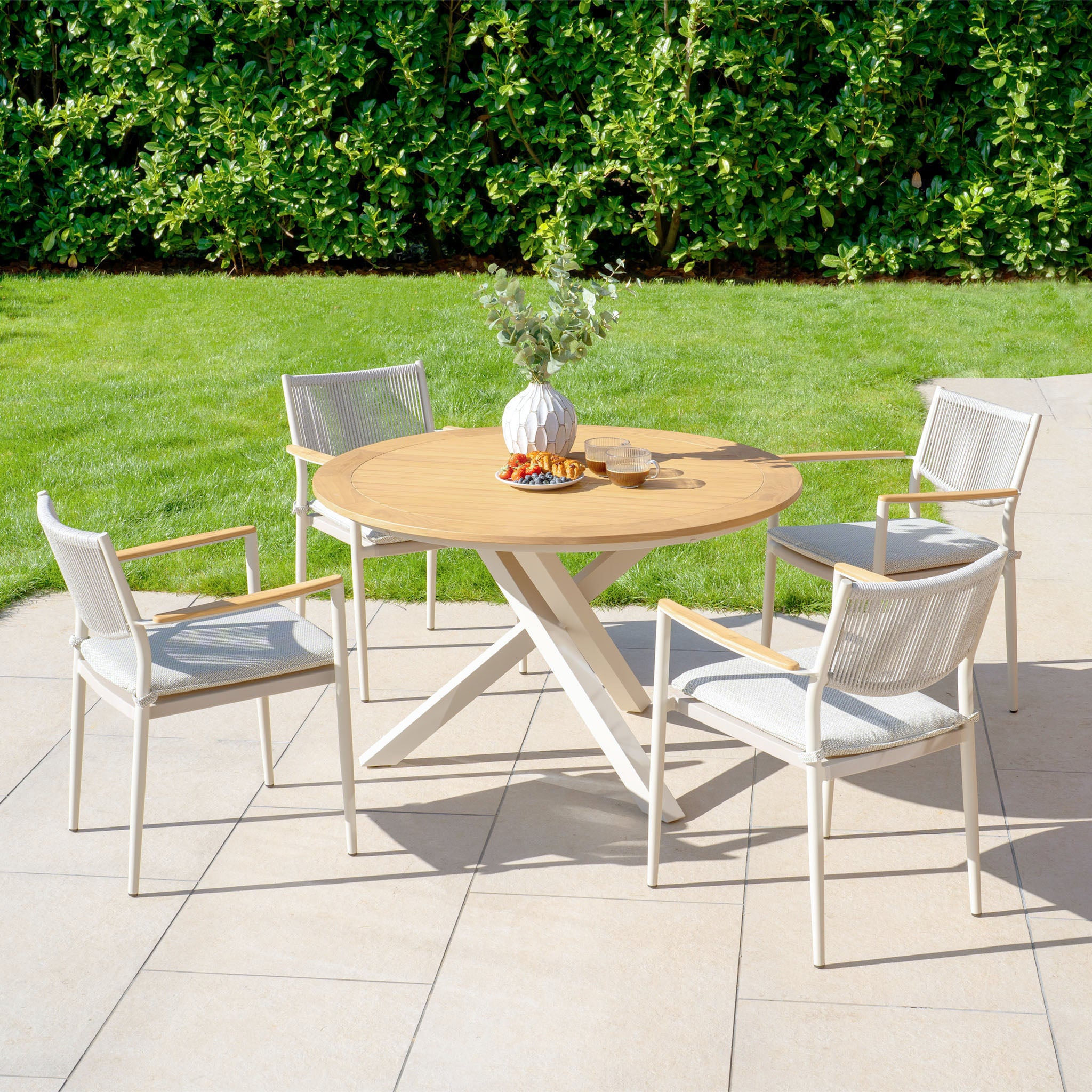 The Tellaro 4 Seat Round Dining Set with Teak Table in Latte boasts a round teak table and four chairs crafted with weather-resistant rope, making it ideal for your patio. It looks stunning on a tiled surface, surrounded by lush green foliage and adorned with a tasteful vase on the table.