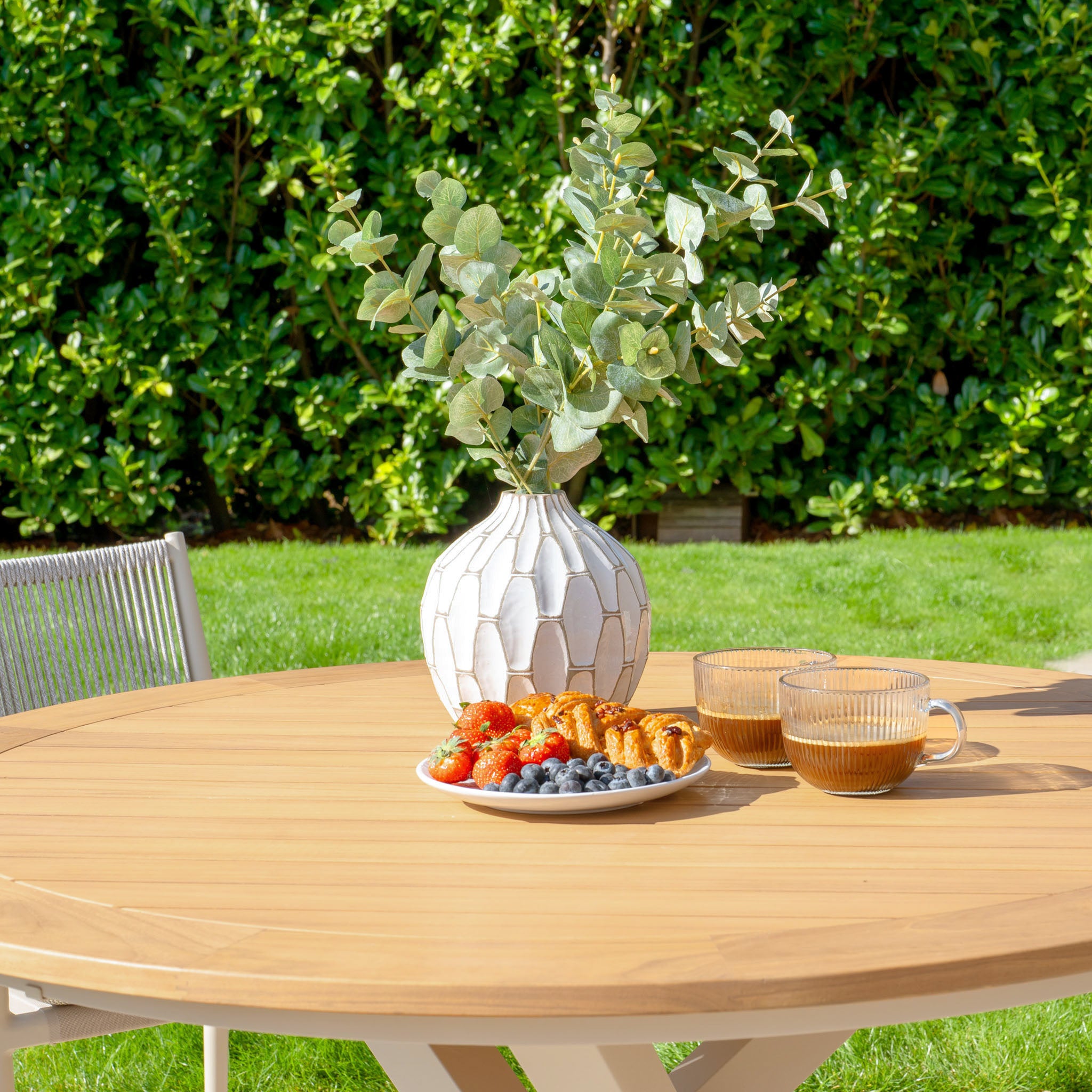 The Tellaro 4 Seat Round Dining Set with a Teak Table in Latte, adorned with coffee, pastries, berries, and a vase of leaves, creates the perfect outdoor dining experience on a sunny day.