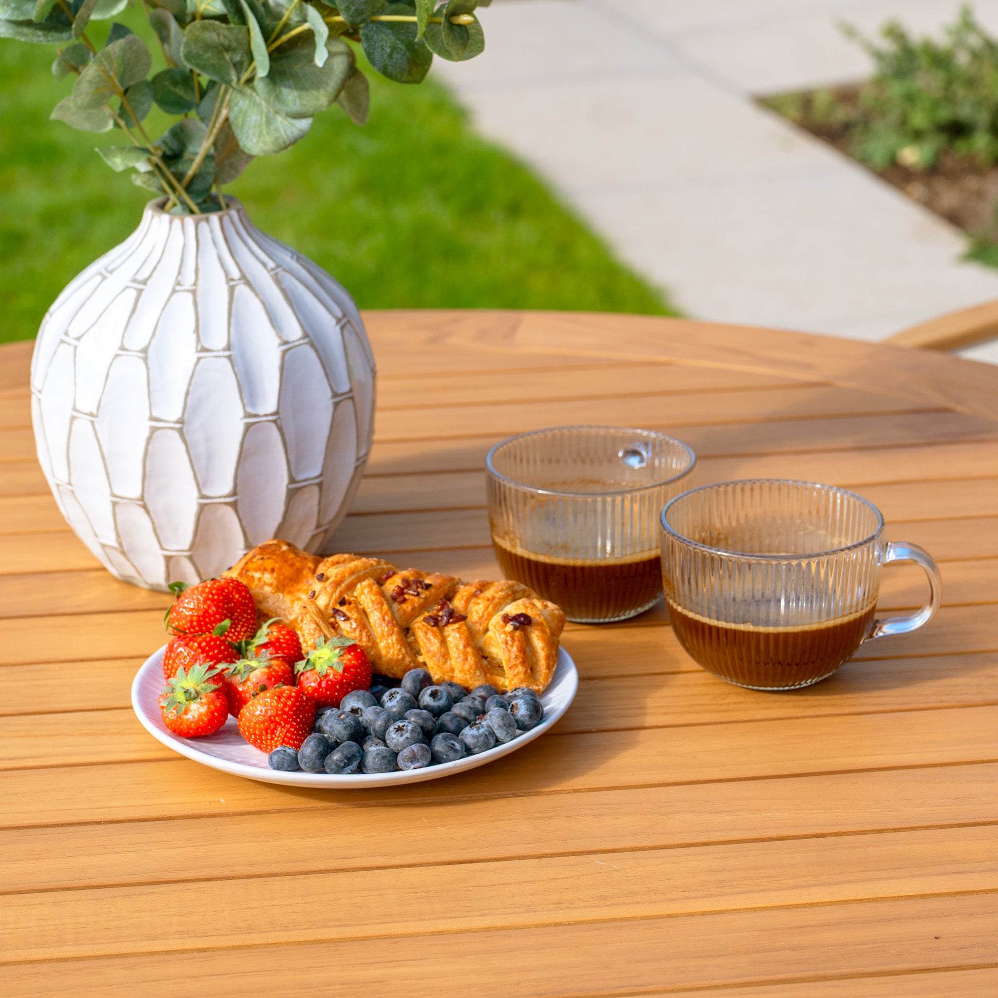 The Tellaro 4 Seat Round Dining Set with a Teak Table in Latte elegantly graces the outdoors, complete with coffee cups, a vase, and a plate of pastries. Strawberries and blueberries add vibrant color, while weather-resistant rope weaves through the chair backs for both style and durability.