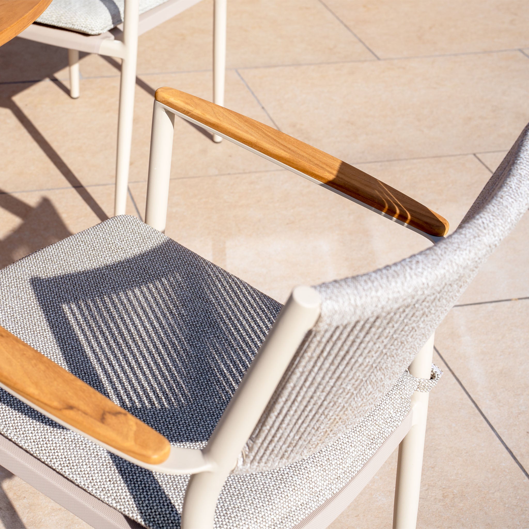 The Tellaro 4 Seat Round Dining Set with a teak table in latte features outdoor chairs with wooden armrests and gray fabric, casting shadows on a light stone patio. This elegant set is ideal for enjoying sunny days in style.