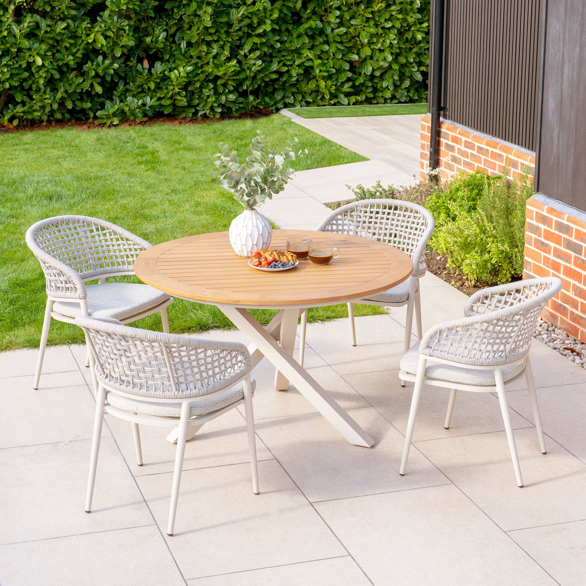 The Kalama 4 Seat Round Dining Set with Teak Table in Latte features a round teak table and four woven chairs, perfect for a stone patio surrounded by lush greenery.