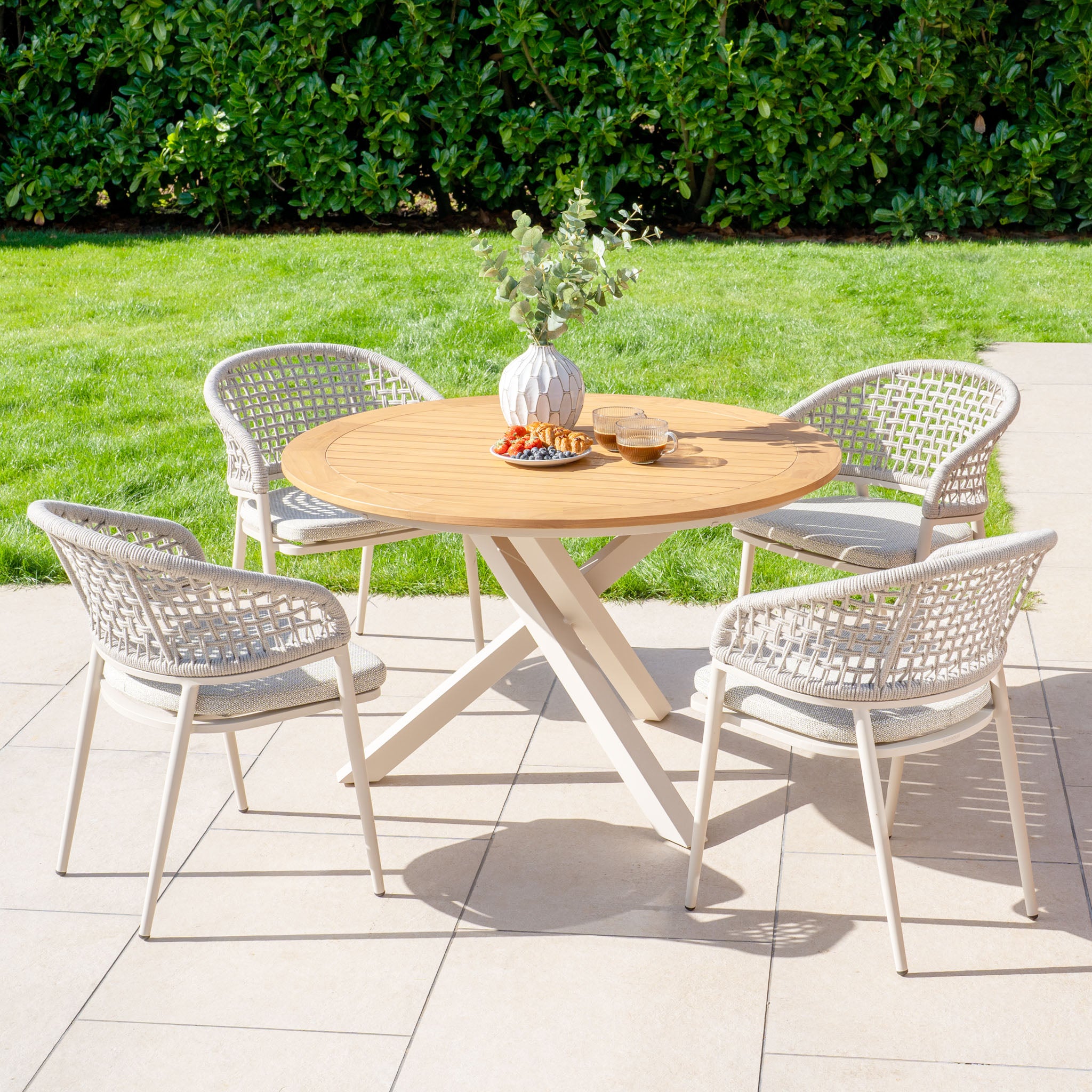 Kalama 4 Seat Round Dining Set with a round teak table in latte, paired with four woven chairs and a decorative vase, placed on a tiled terrace near the lawn.