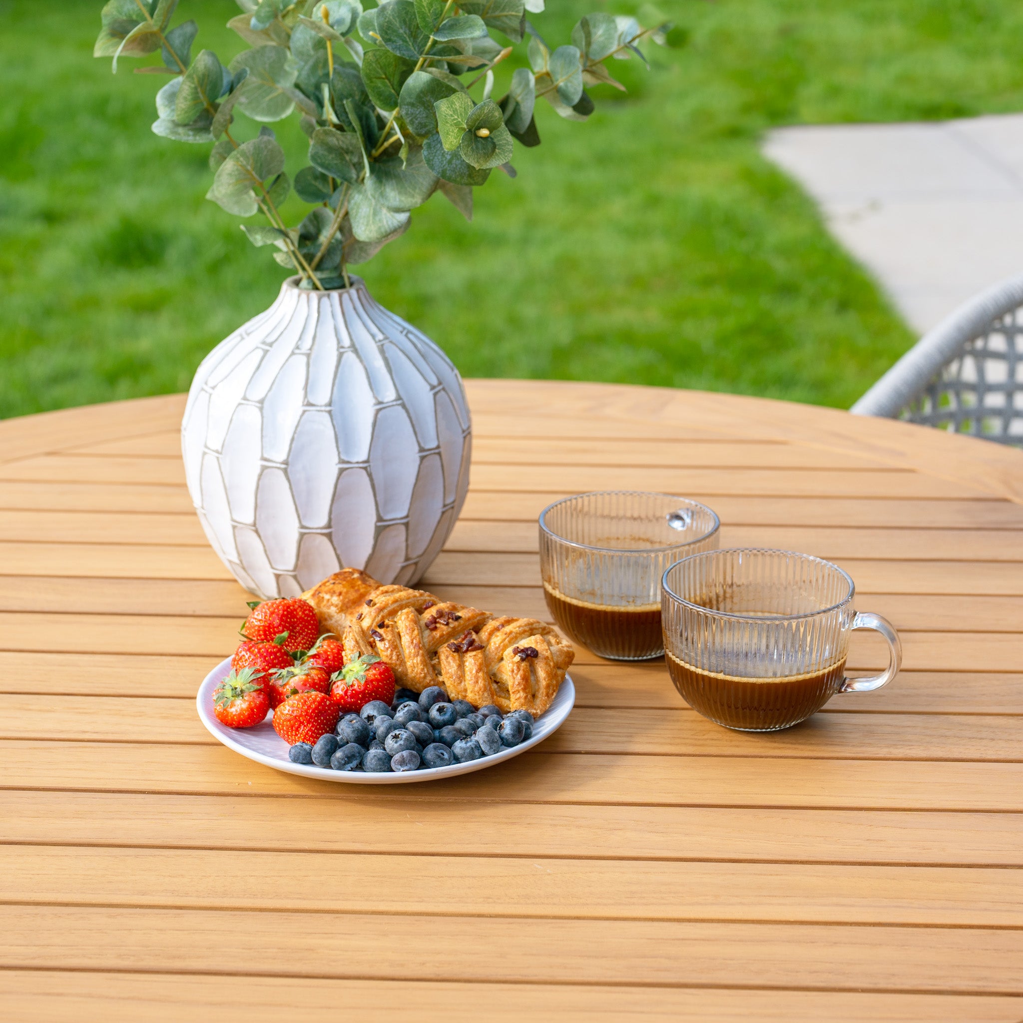 The Kalama 4 Seat Round Dining Set, featuring a teak table in latte, holds coffee and pastries with strawberries and blueberries elegantly displayed on its rustic surface. The woven chairs from the outdoor dining set create a perfect setting for enjoying the fresh air.