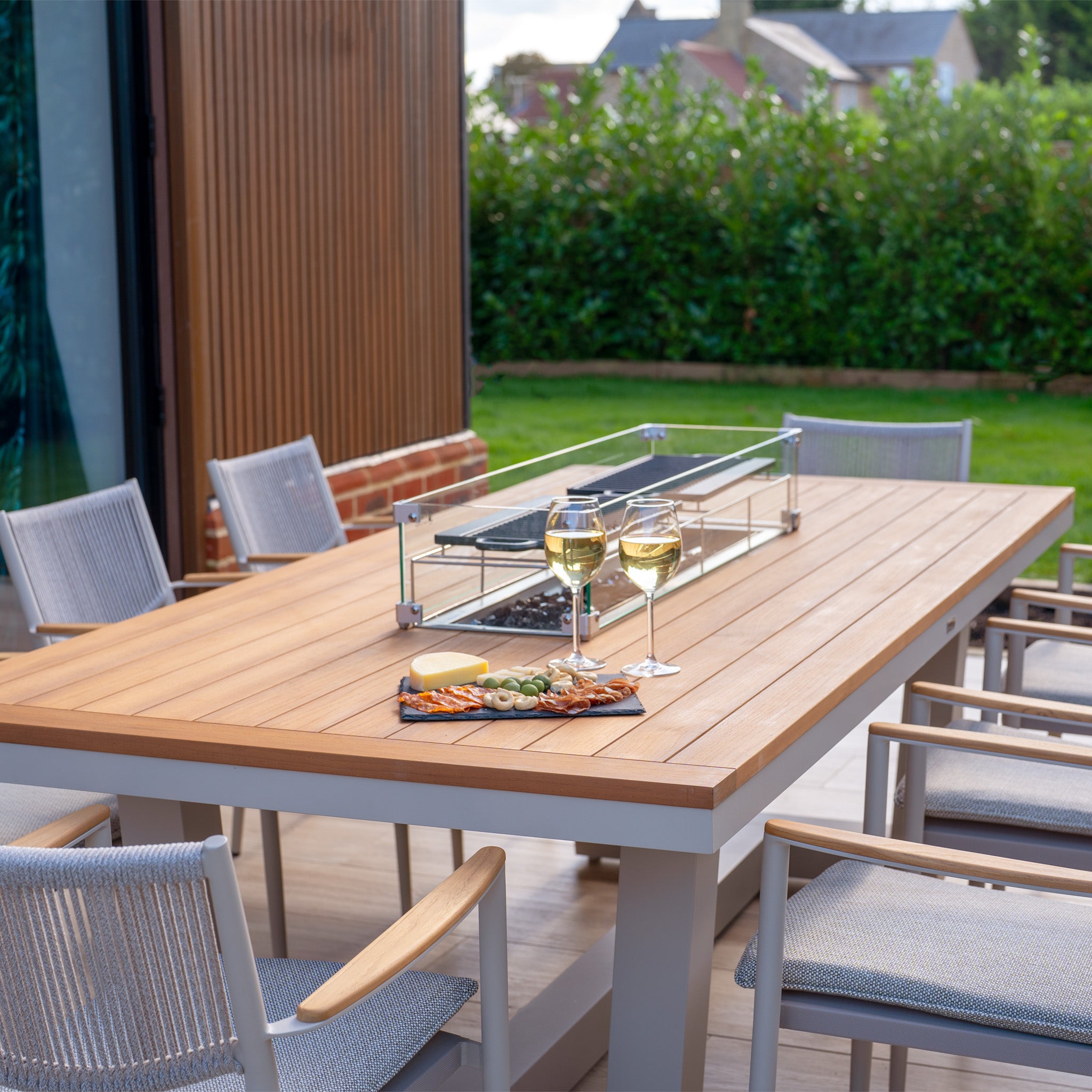 The patio showcases a beautiful outdoor dining scene with the Tellaro 8 Seat Rectangular Teak Firepit Dining Set in Latte, accompanied by a charcuterie board and two glasses of white wine, perfectly accented by the stylish Tellaro Chairs.