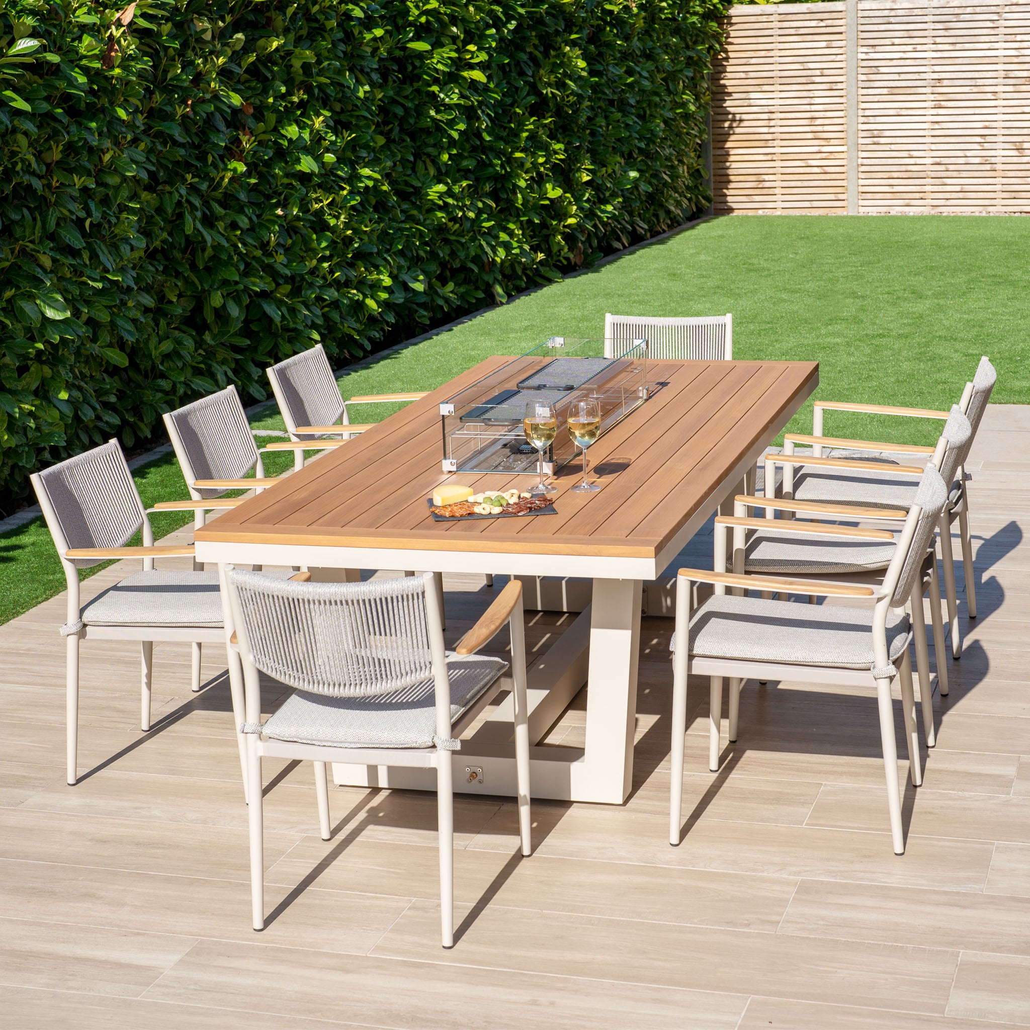 The outdoor patio showcases a spacious Tellaro 8 Seat Rectangular Teak Firepit Dining Set in Latte, ideal for gatherings. Surrounded by lush greenery and grass, it invites relaxation while keeping the ambiance warm and welcoming.