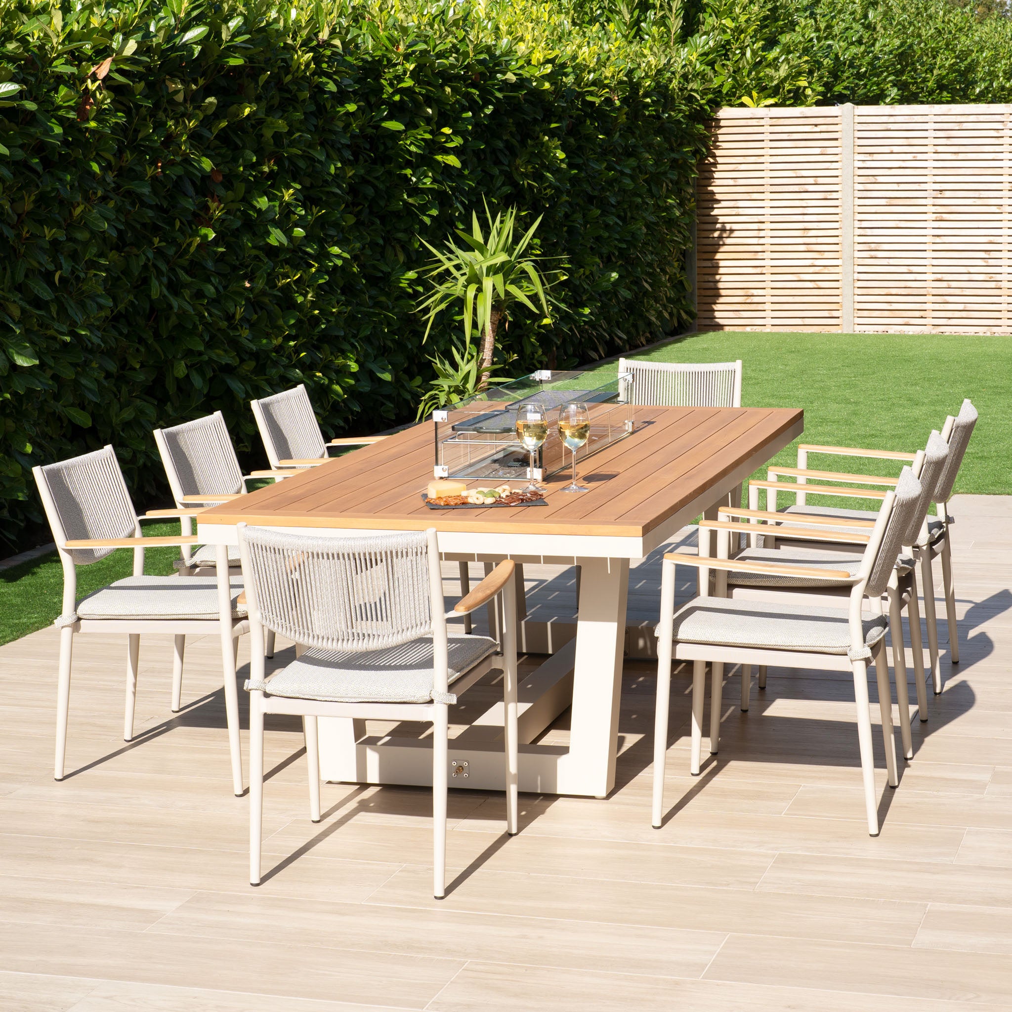 Host delightful outdoor meals with our Tellaro 8 Seat Rectangular Teak Firepit Dining Set in Latte, featuring a wooden table and eight matching chairs. Ideal for a patio surrounded by lush greenery, this setup is perfect for cozy gatherings around the firepit with drinks and friends.