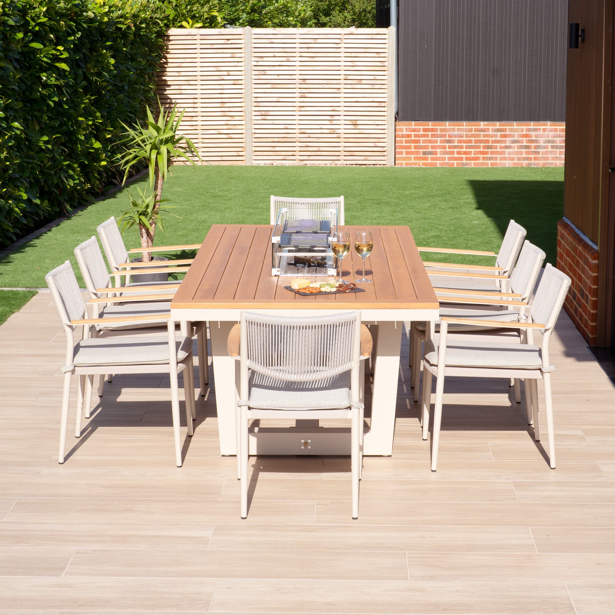 An outdoor patio showcases the Tellaro 8 Seat Rectangular Teak Firepit Dining Set in Latte, featuring a wooden table and eight matching chairs. Two wine glasses accompany a plate of snacks, creating a picturesque scene against a lush grassy backdrop.