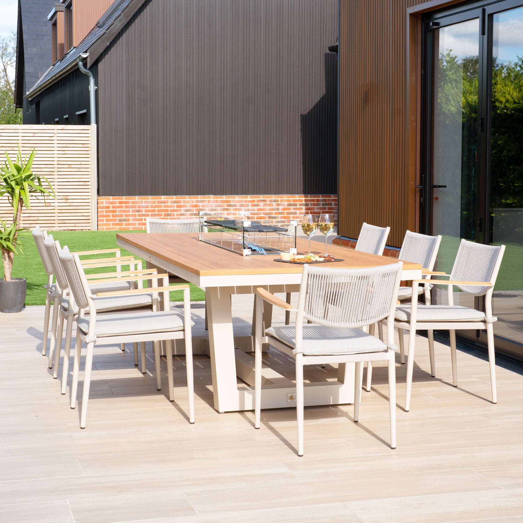 The patio is adorned with a Tellaro 8 Seat Rectangular Teak Firepit Dining Set in Latte, featuring sleek chairs and an outdoor table highlighted by powder-coated aluminium accents, all elegantly set with drinks and snacks to perfectly complement the modern house.