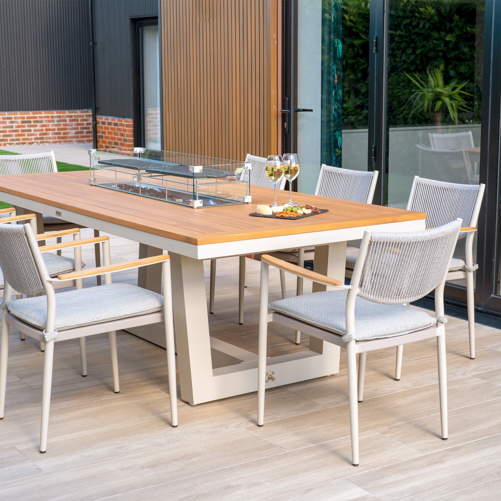 Gather your friends around the Tellaro 8 Seat Rectangular Teak Firepit Dining Set in Latte, featuring a wooden dining table paired with matching chairs, wine glasses, and a snack tray on a modern wooden deck. Perfect for cozy evenings under the stars.