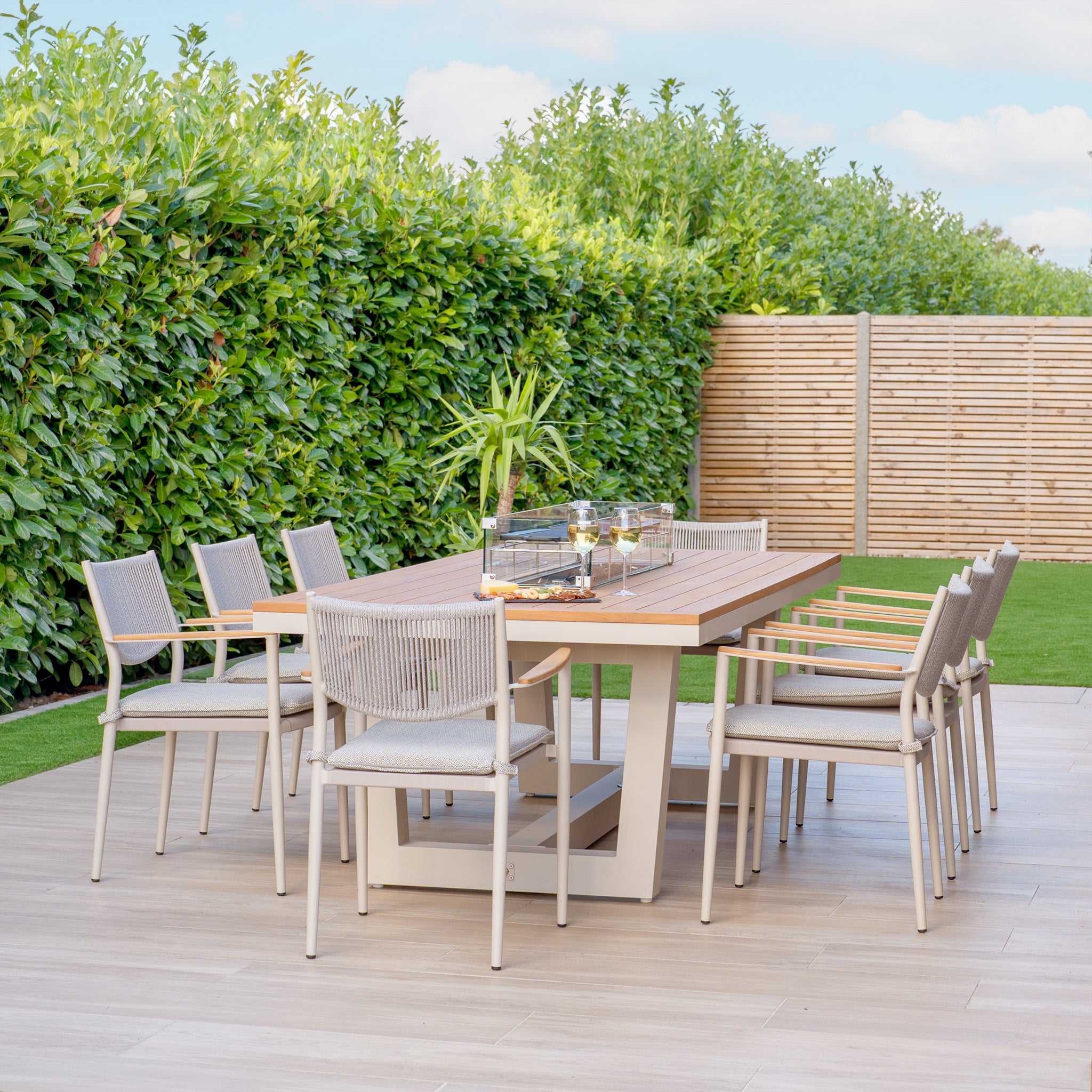 The inviting outdoor dining area features a wooden table paired with chairs, all arranged on a cozy patio. A small plant centerpiece adds charm, while the setting gains warmth and elegance from the Tellaro 8 Seat Rectangular Teak Firepit Dining Set in Latte and nearby hedges.