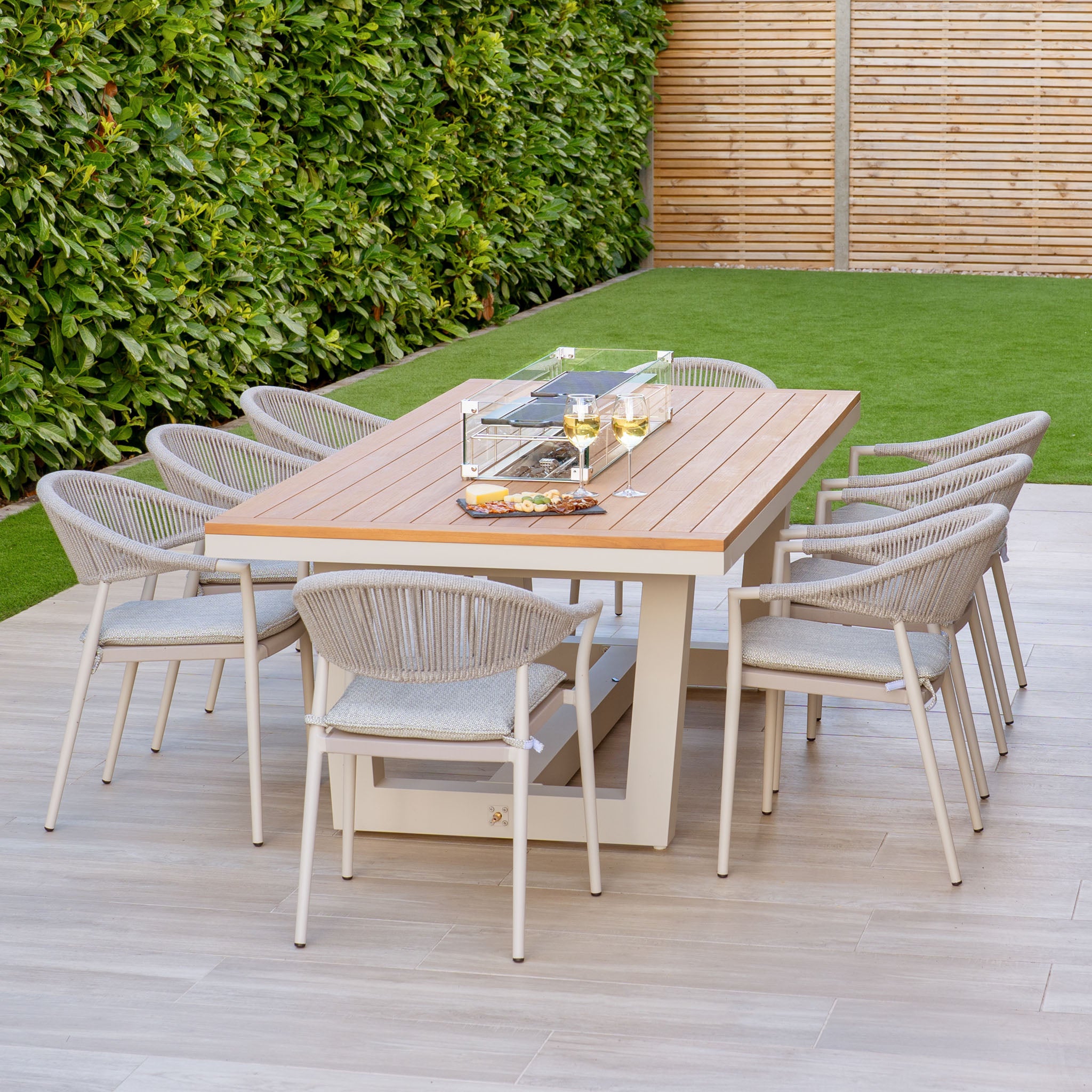 The Cloverly 8 Seat Rectangular Teak Firepit Dining Set in Latte includes a wooden table surrounded by eight UV-resistant chairs and a tabletop grill, all beautifully situated among lush green foliage.