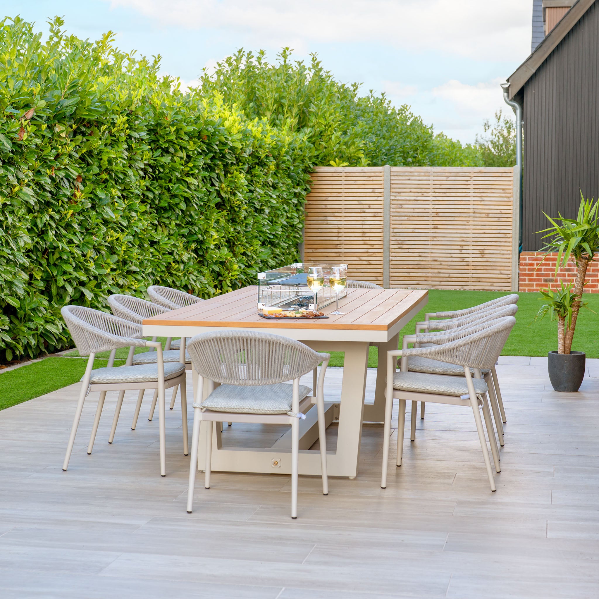 The stylish Cloverly 8 Seat Rectangular Teak Firepit Dining Set in Latte enhances the patio, bordered by vibrant green hedges and a potted plant. The aluminum components offer a sleek appearance, while the UV-resistant chairs ensure enduring comfort as delightful snacks adorn the table.