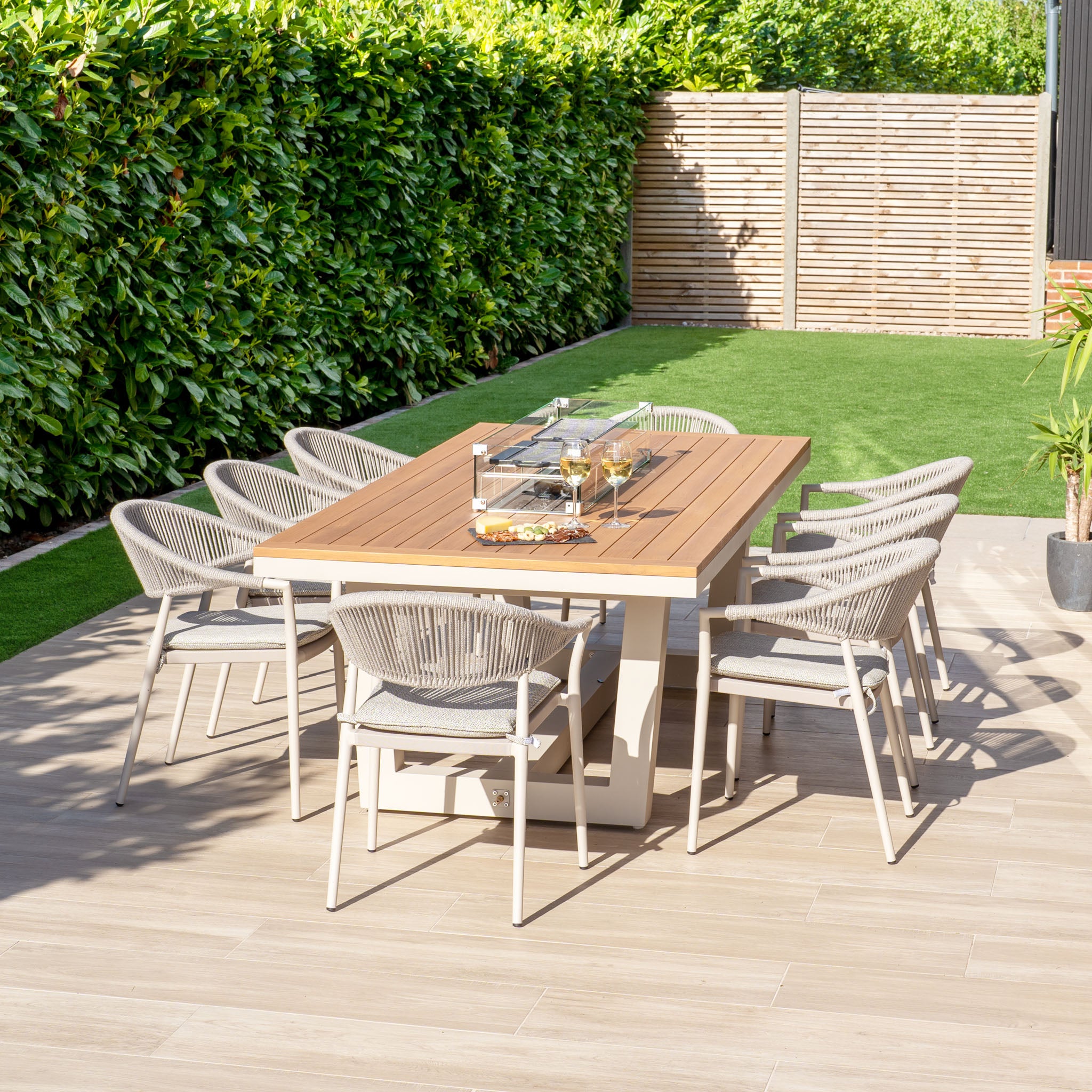 The Cloverly 8 Seat Rectangular Teak Firepit Dining Set in Latte showcases a stunning wooden table and eight UV-resistant chairs, elegantly positioned on a patio amid lush greenery and encircled by a wooden fence.